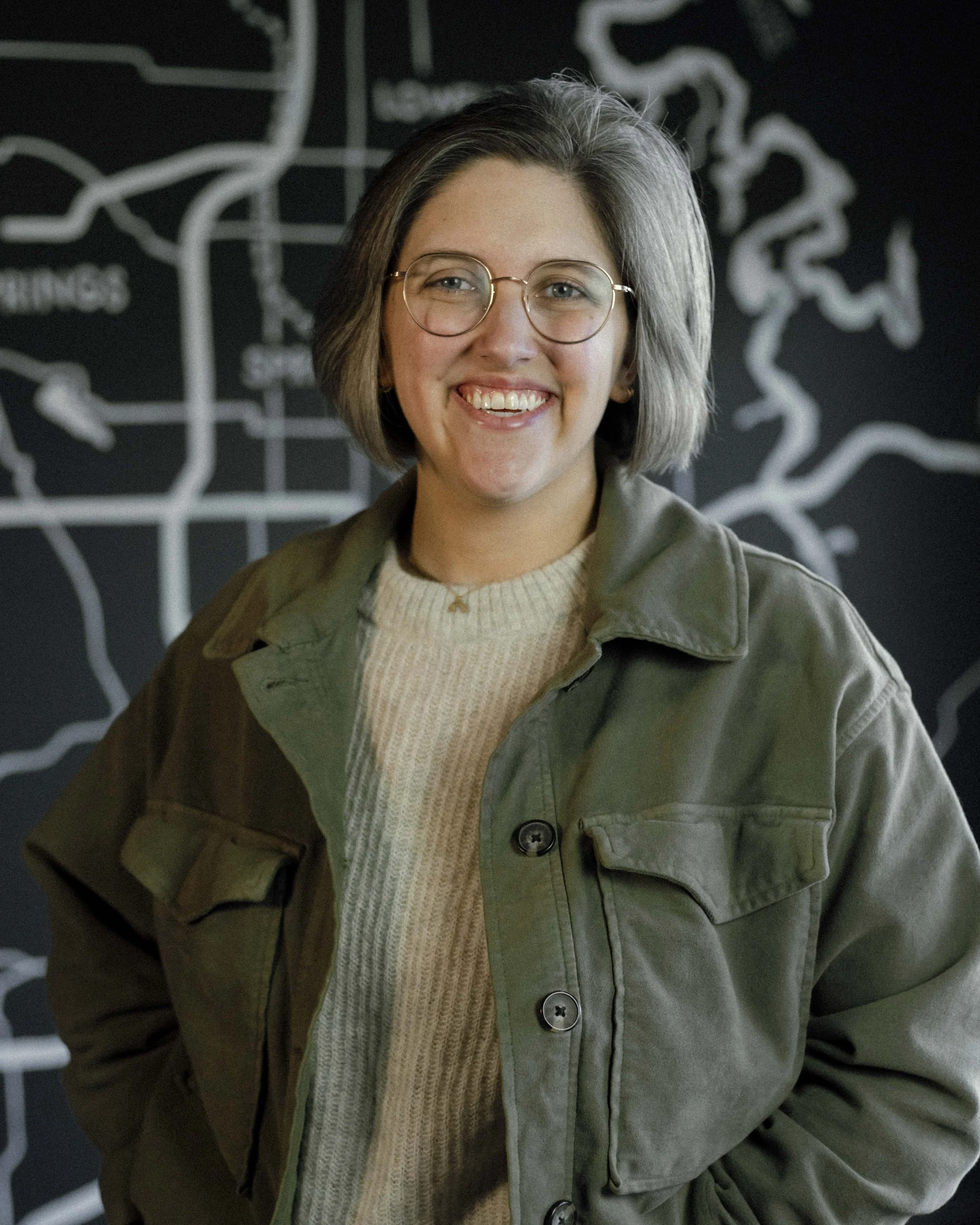 A woman with short gray hair, glasses, and a big smile wearing a beige sweater and an olive green jacket, standing in front of a black chalkboard with white chalk drawings of a map.