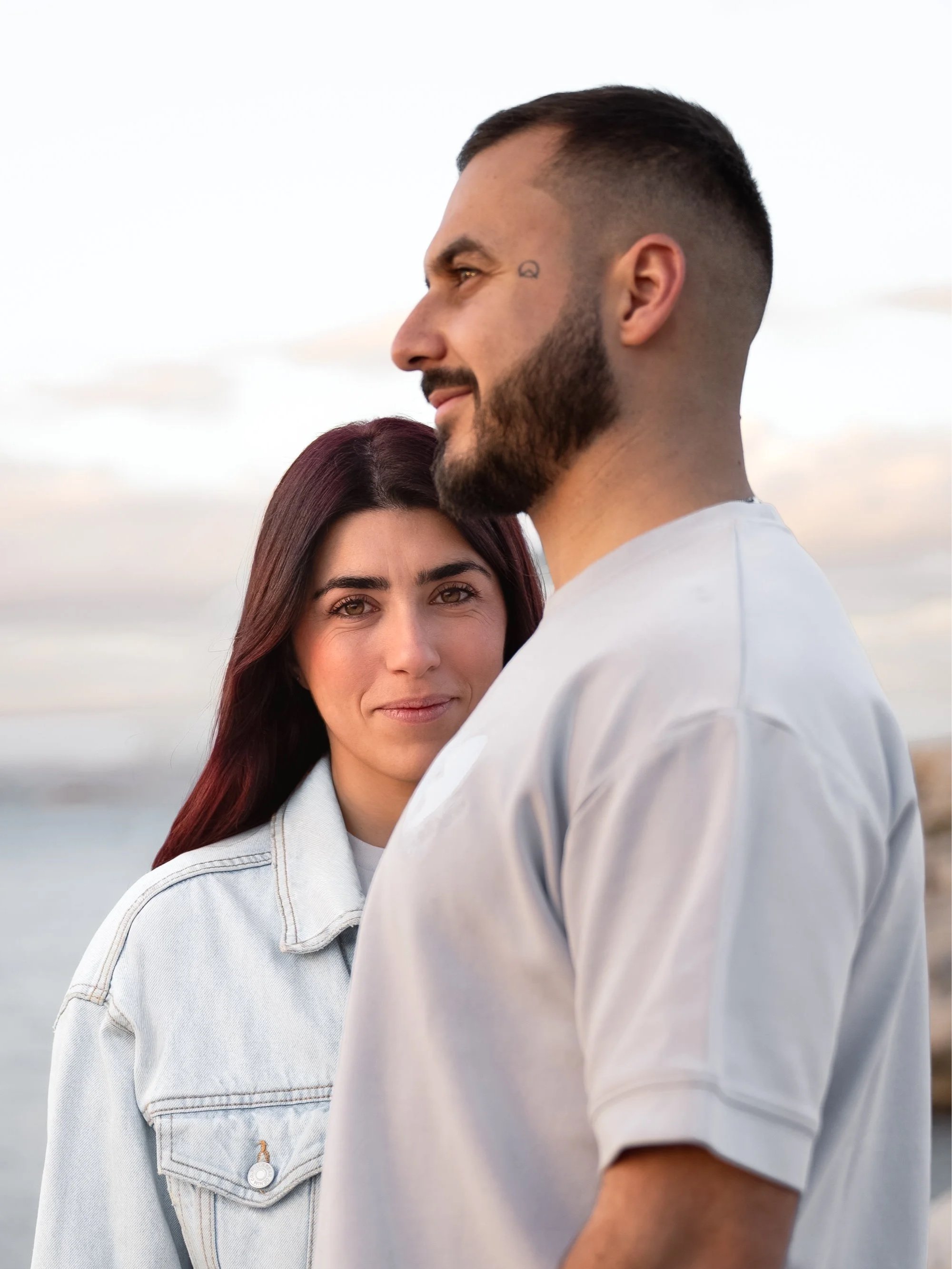 Un couple regarde l'objectif avec la mer en arrière-plan, au coucher du soleil, dans une ambiance romantique.