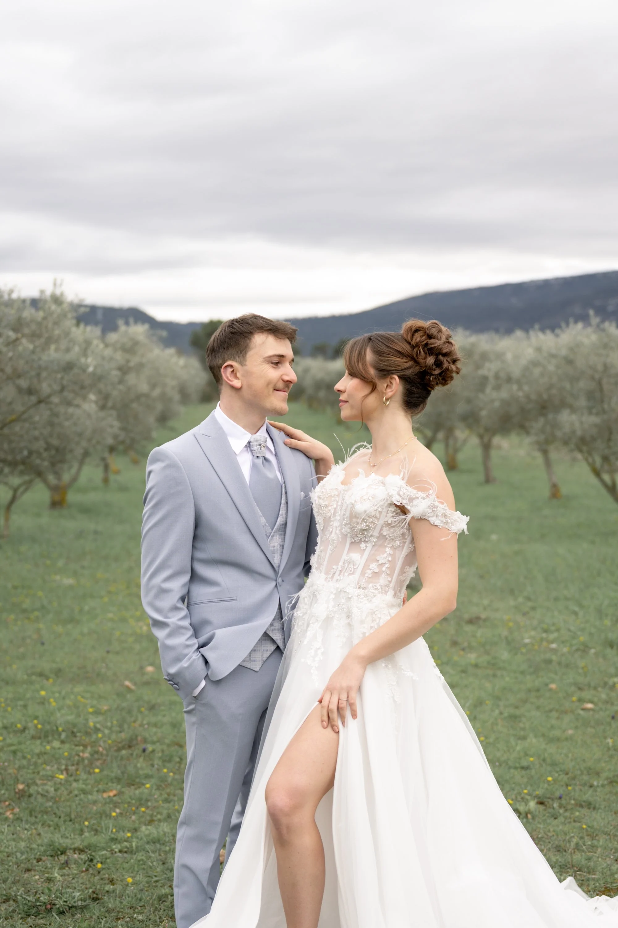 Un couple de mariés en costume et robe de mariage se regarde dans un champ avec des arbres et des montagnes en arrière-plan.