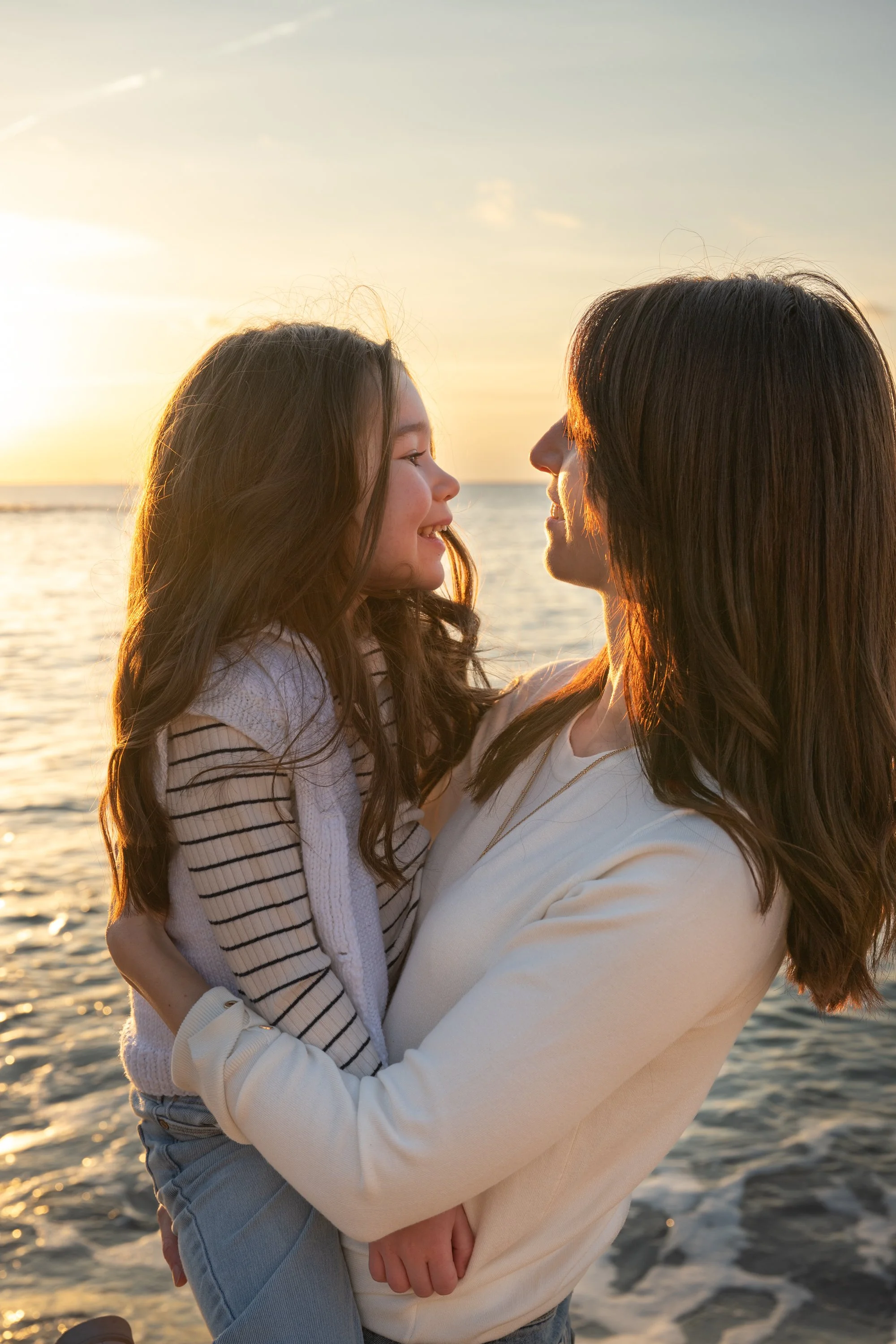 Une mère et sa fille souriantes se regardent devant la mer au coucher du soleil.