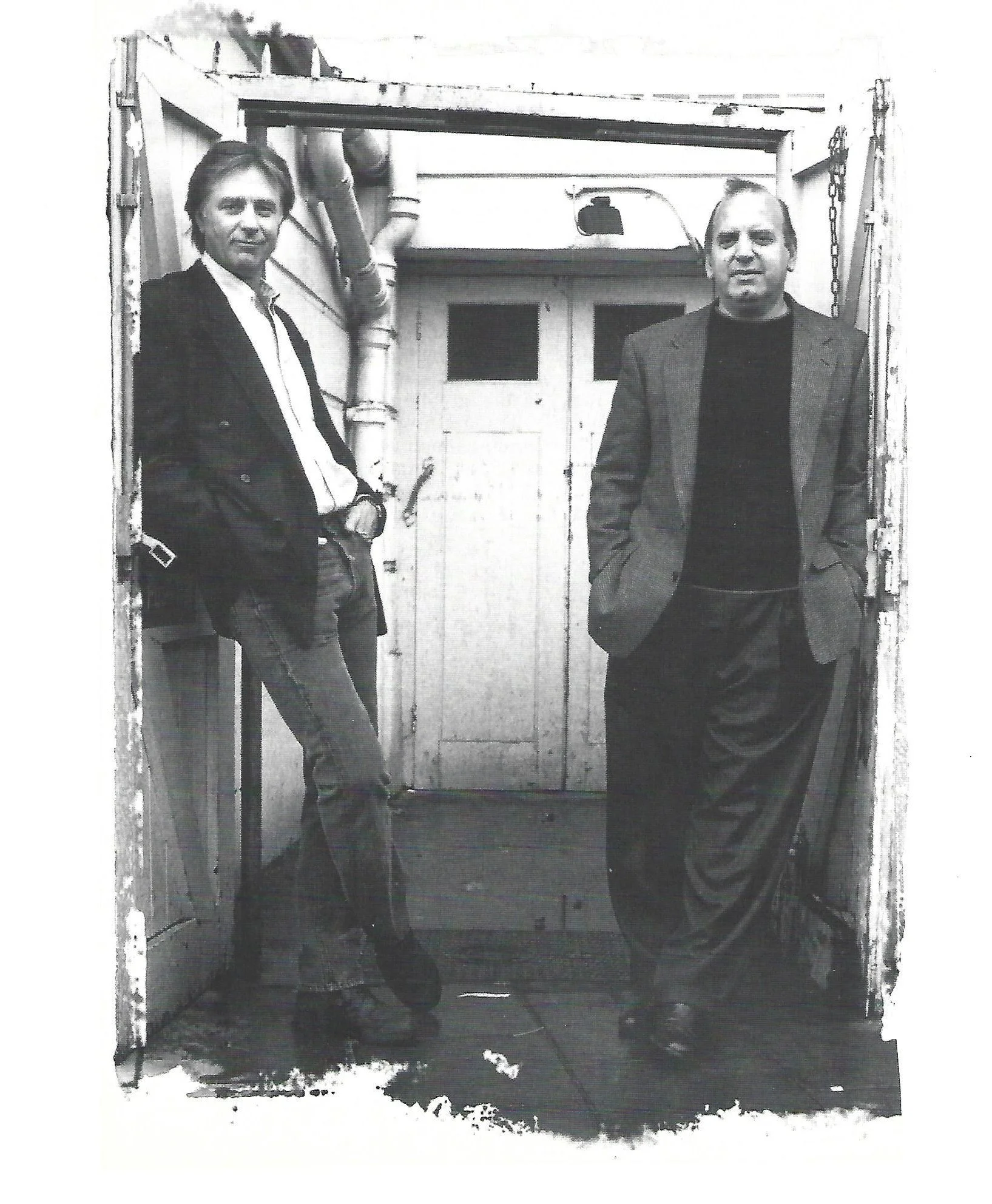 Two men standing inside a doorway, both facing forward. The man on the left has dark hair, wears a dark blazer over a white shirt, has a beard, and is smiling slightly. The man on the right has lighter hair, wears a darker blazer over a dark shirt, has a beard, and looks serious. There are some pipes and equipment visible in the background.