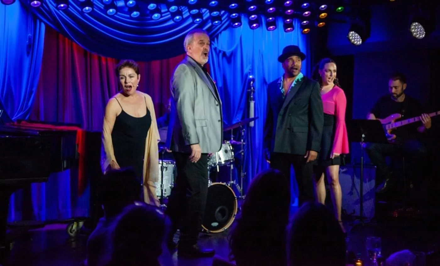 A group of five performers on a stage with colorful blue and purple curtains, singing and playing music. One woman in a black dress and yellow cardigan, two men in suits, one woman in a pink dress, and a guitarist in black, with drums in the backgrou