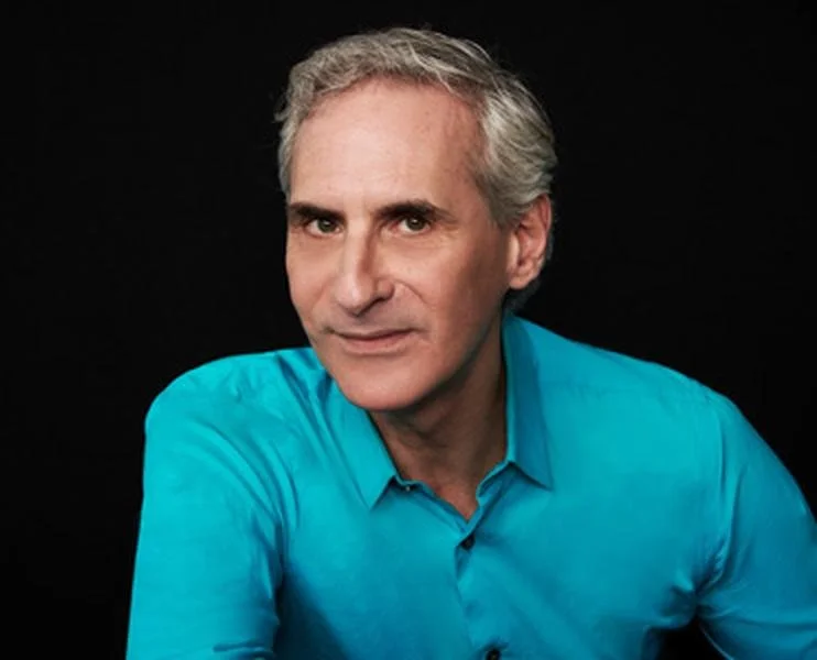 A middle-aged man with gray hair wearing a turquoise shirt, posing against a black background.