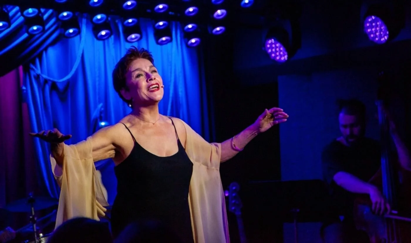 A woman singing on stage with her arms outstretched, in front of a blue curtain and stage lights, with a pianist in the background.