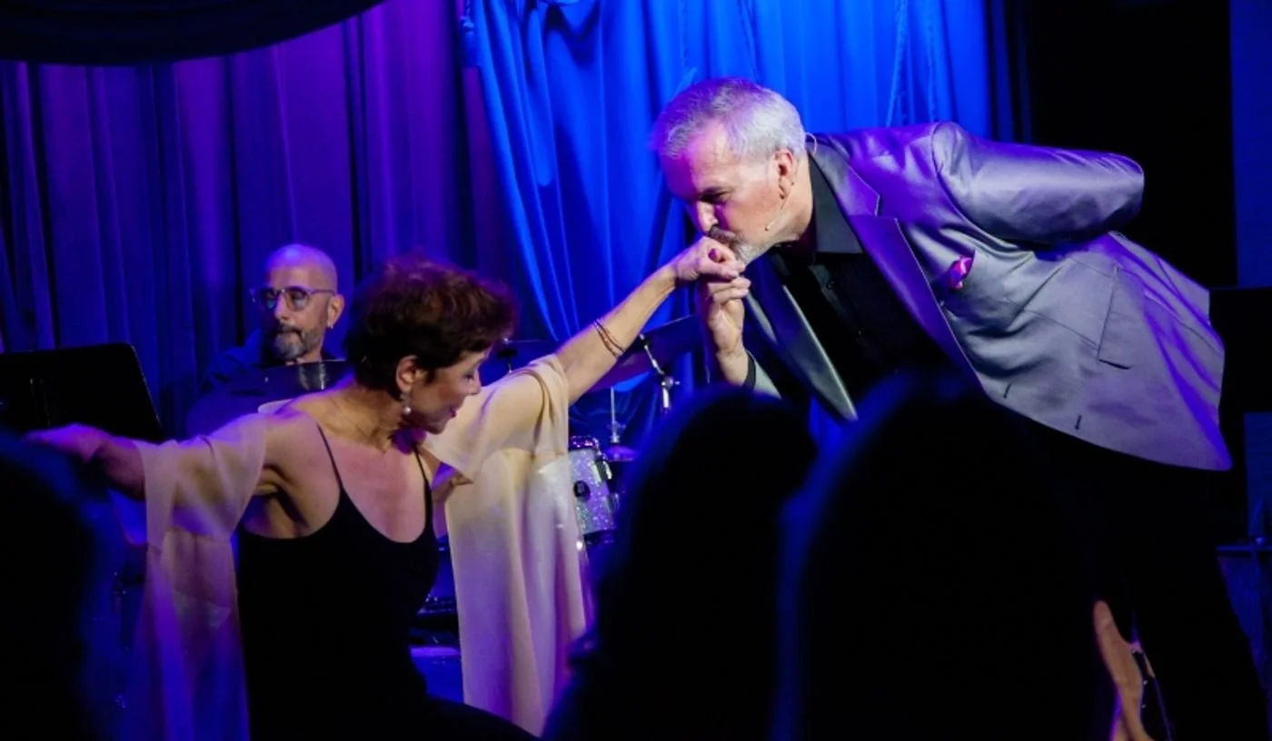 A man in a gray blazer is kissing the hand of a woman in a black dress on stage with a blue curtain backdrop, while another man sits in the background.