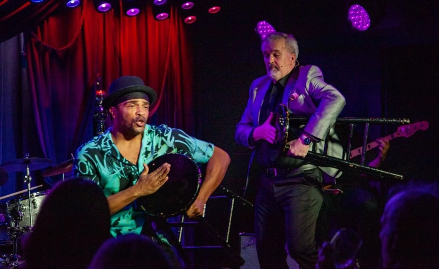 Two musicians performing on a stage with red curtains, one playing percussion and singing, the other holding a bass guitar, under purple lighting, with an audience visible in the foreground.