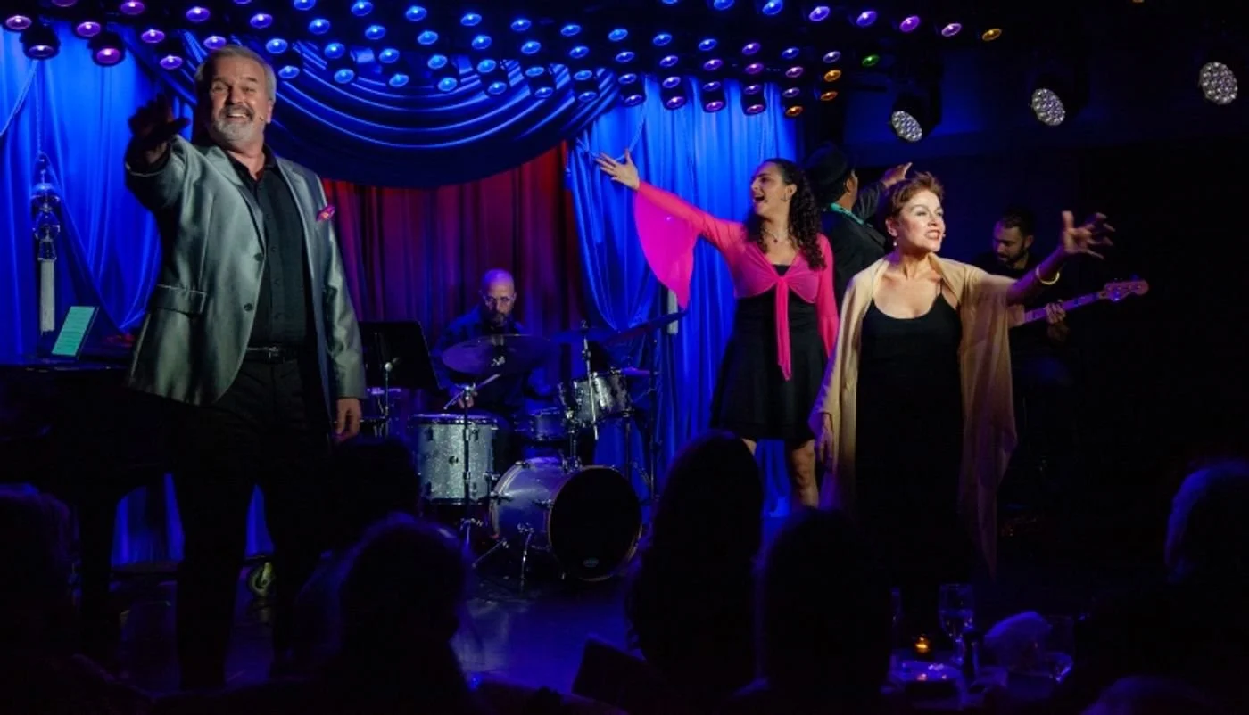 Group of five performers on stage with a drummer, two women and two men, under stage lights and blue curtains, performing or singing with audience visible in foreground.