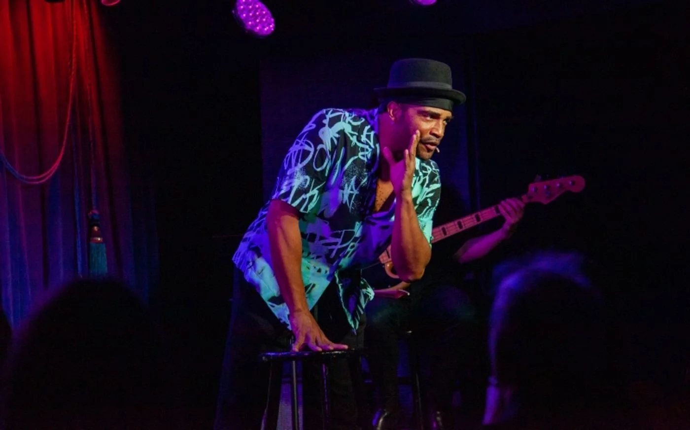 A man in a patterned shirt and fedora hat performing on stage with a microphone, with a bass guitarist partially visible in the background, under purple stage lighting.