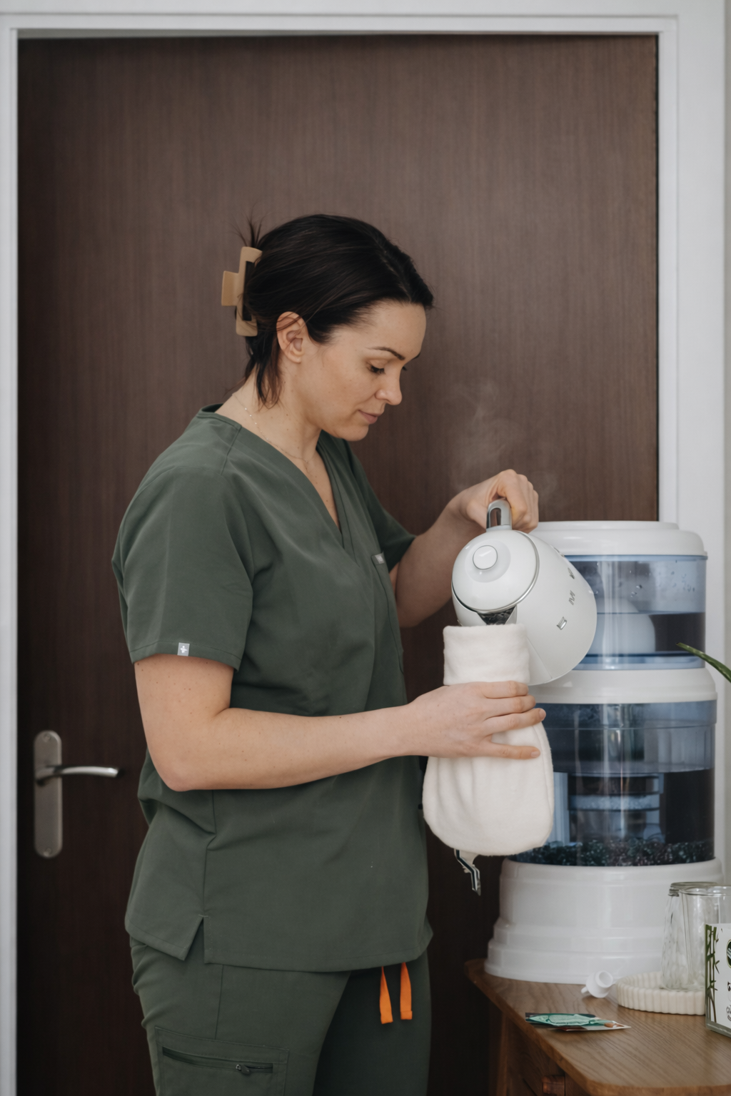 A woman in green medical scrubs is filling a sock with water from a white humidifier.