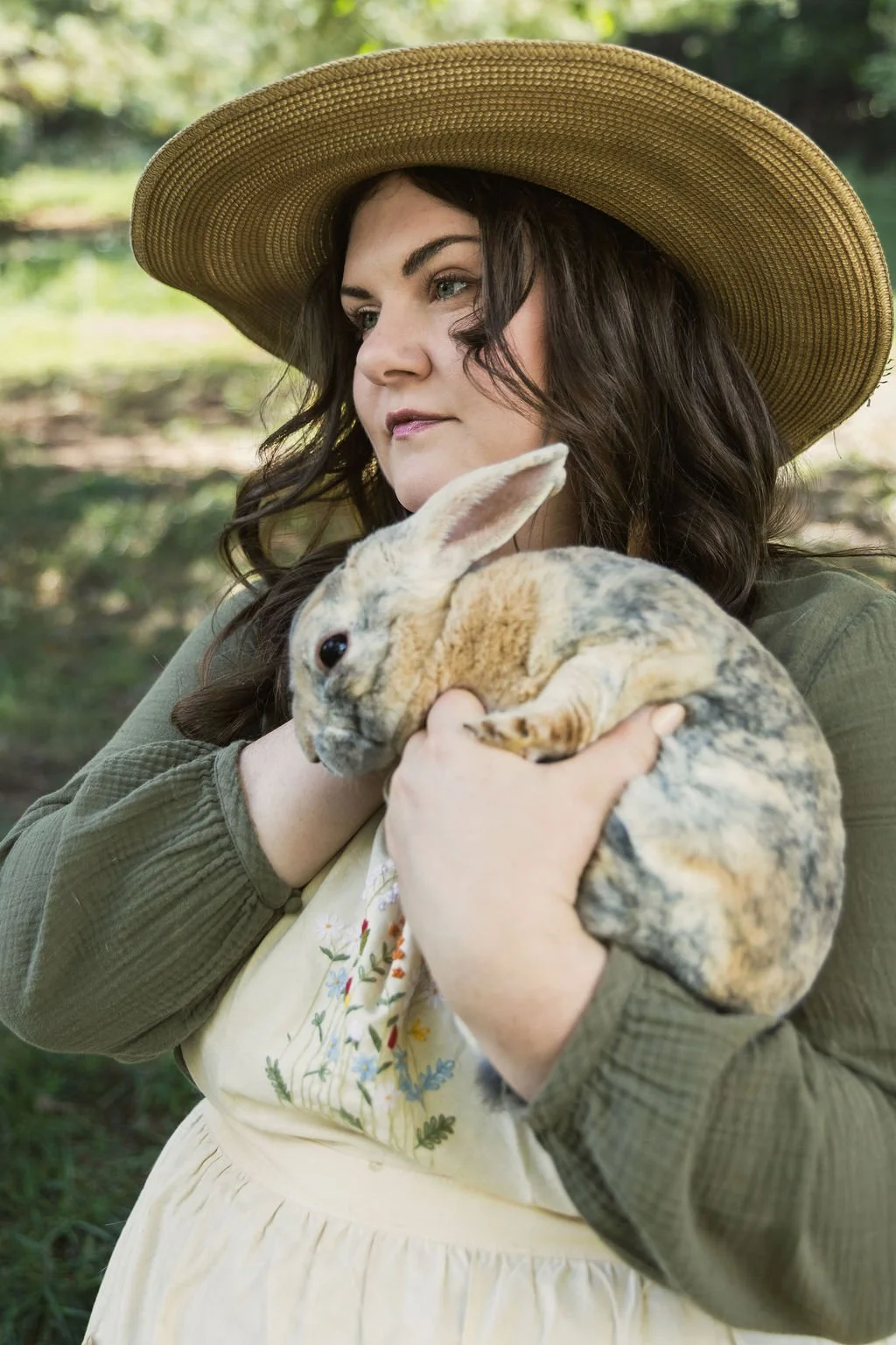 Woman with long dark hair wearing a large straw hat, holding a rabbit outdoors.