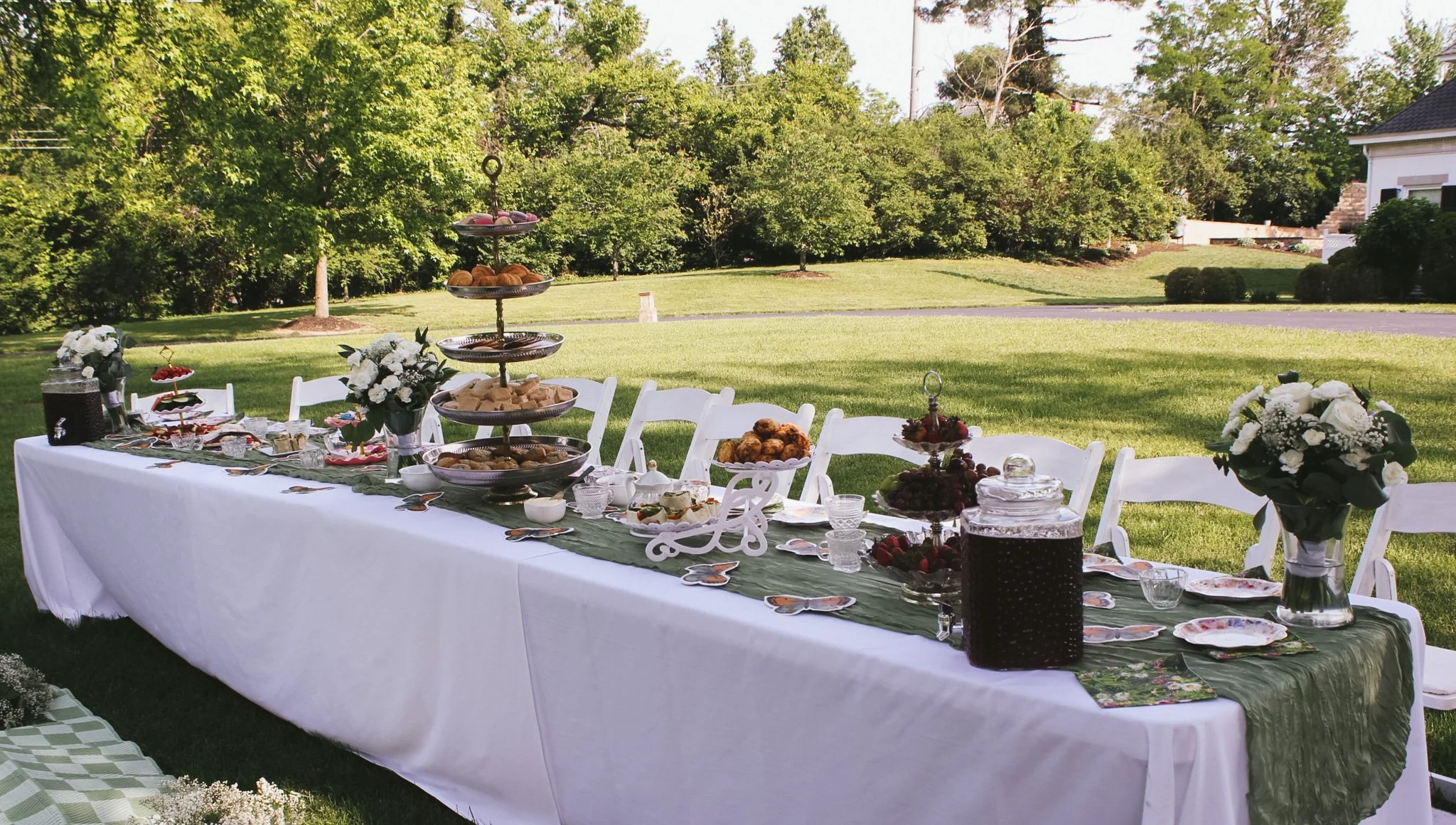 Outdoor tea party event setup with a long table covered in a white tablecloth, decorated with floral arrangements and assorted desserts, set on a grassy lawn with trees in the background.