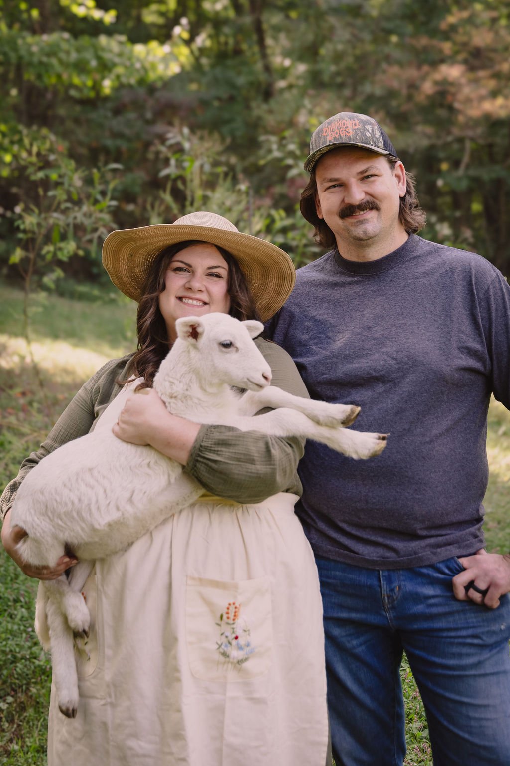 A woman holding a lamb and smiling with a man standing beside her outdoors in a wooded area.
