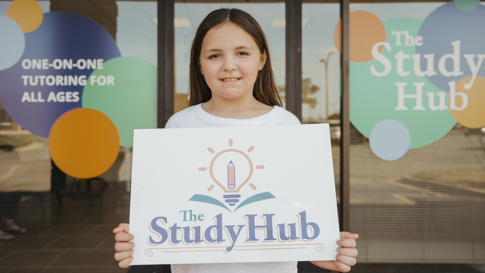 A young girl with brown hair holding a sign that reads "The Study Hub" with an illustration of a lightbulb and pencil above an open book, standing in front of a window with promotional stickers.