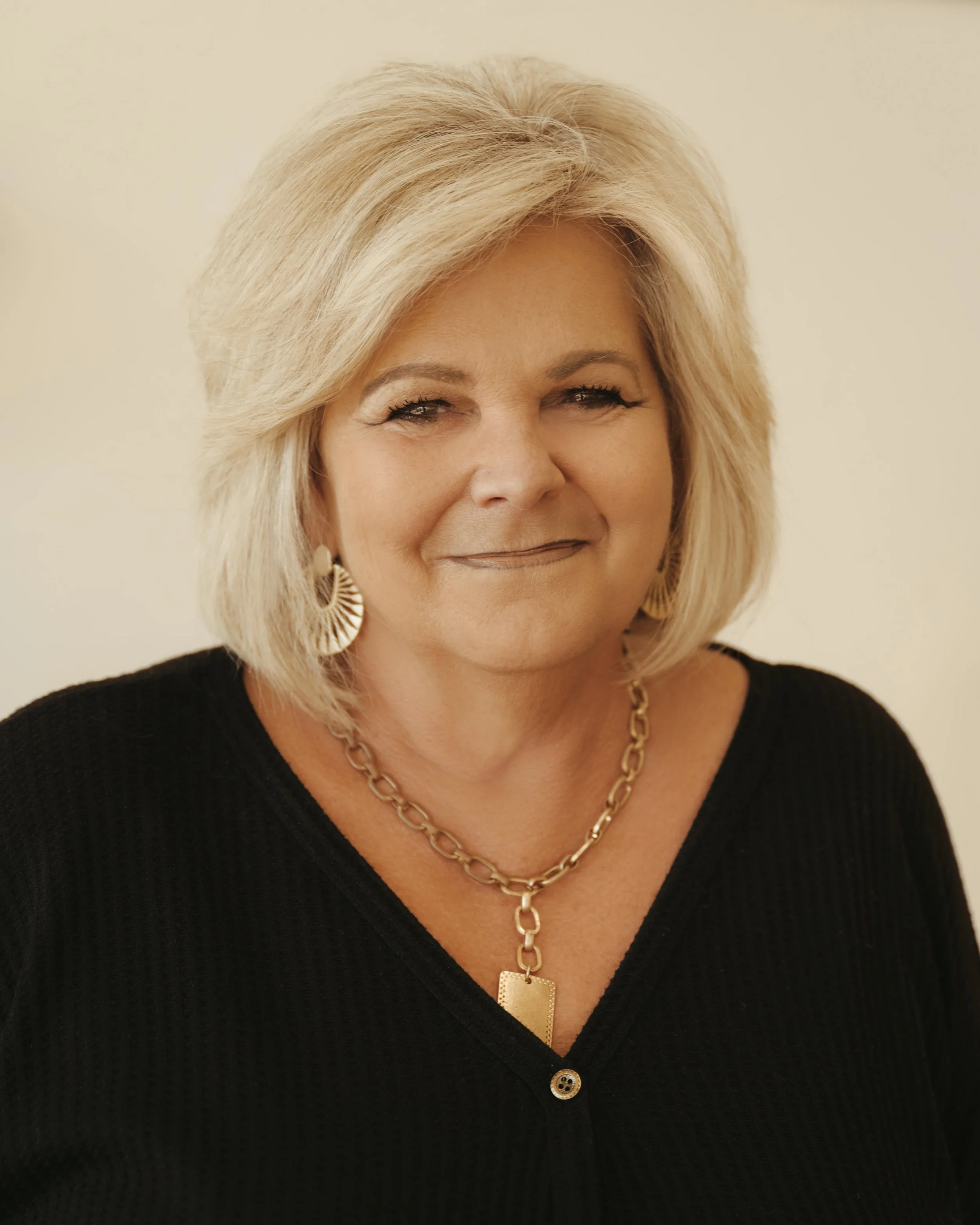 A portrait of a middle-aged woman with blonde hair styled in a layered bob, wearing gold earrings and a chunky gold chain necklace with a rectangular pendant, dressed in a black top, against a neutral background.