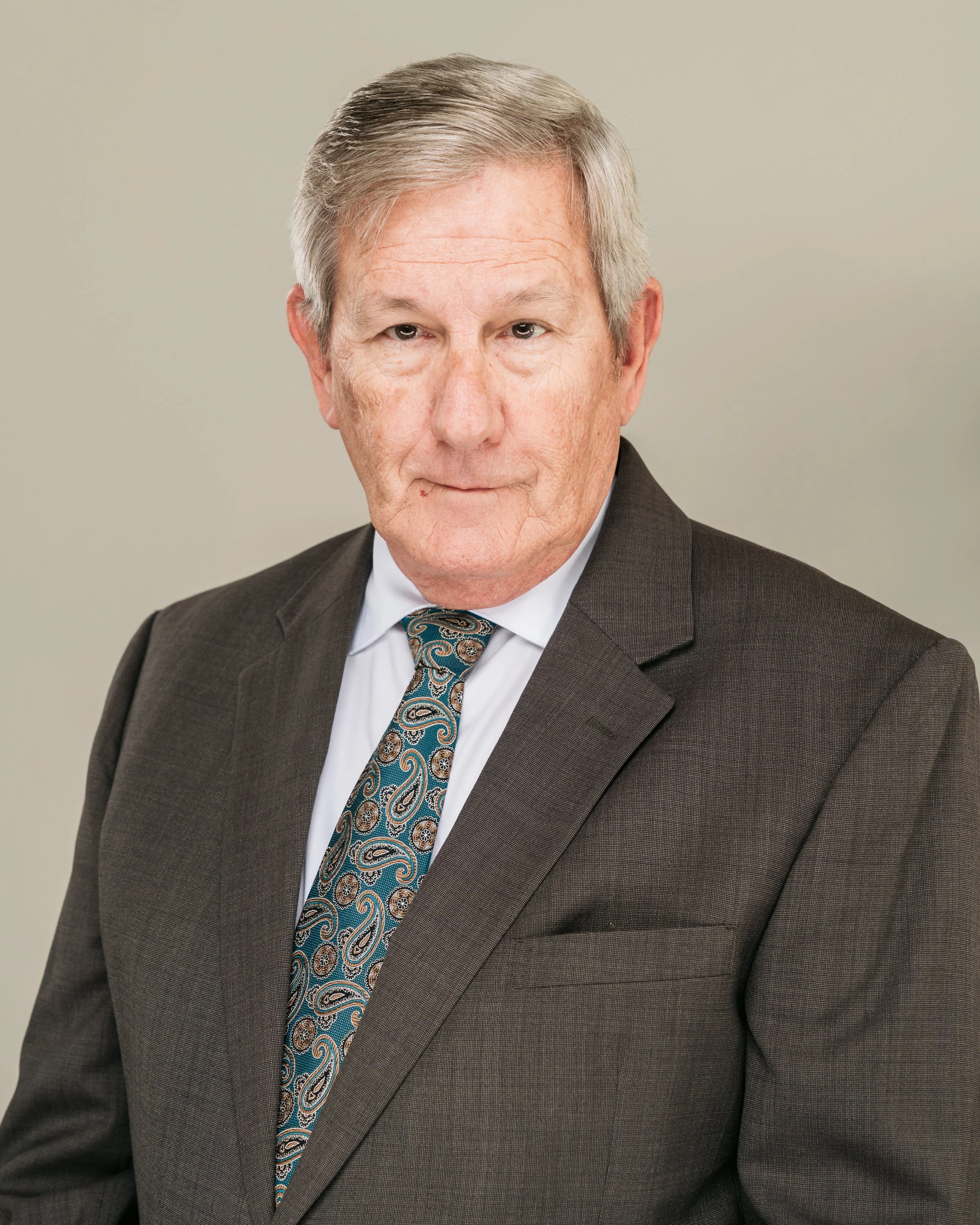 A portrait of an older man wearing a dark suit, white shirt, and paisley tie, with gray hair, looking at the camera against a plain, light-colored background.