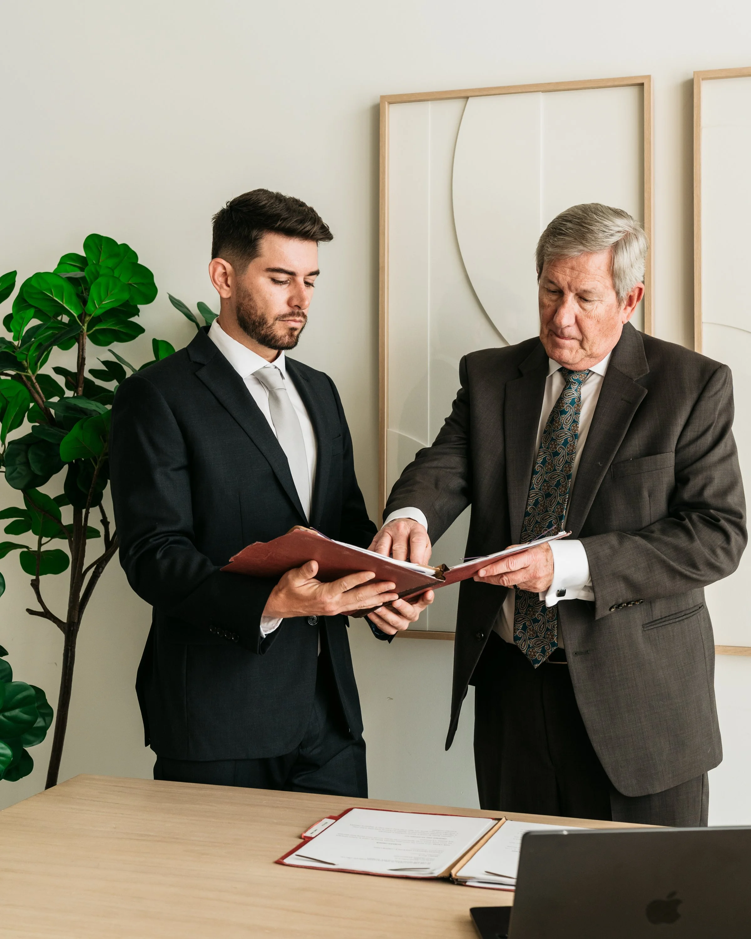 Two men in formal business suits shaking hands over a folder in an office setting, with documents and a laptop on the table.