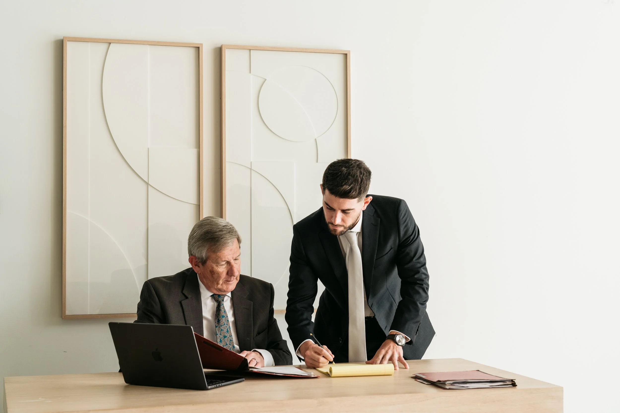 Two men in suits are reviewing documents at a meeting in an office with abstract wall art in the background.