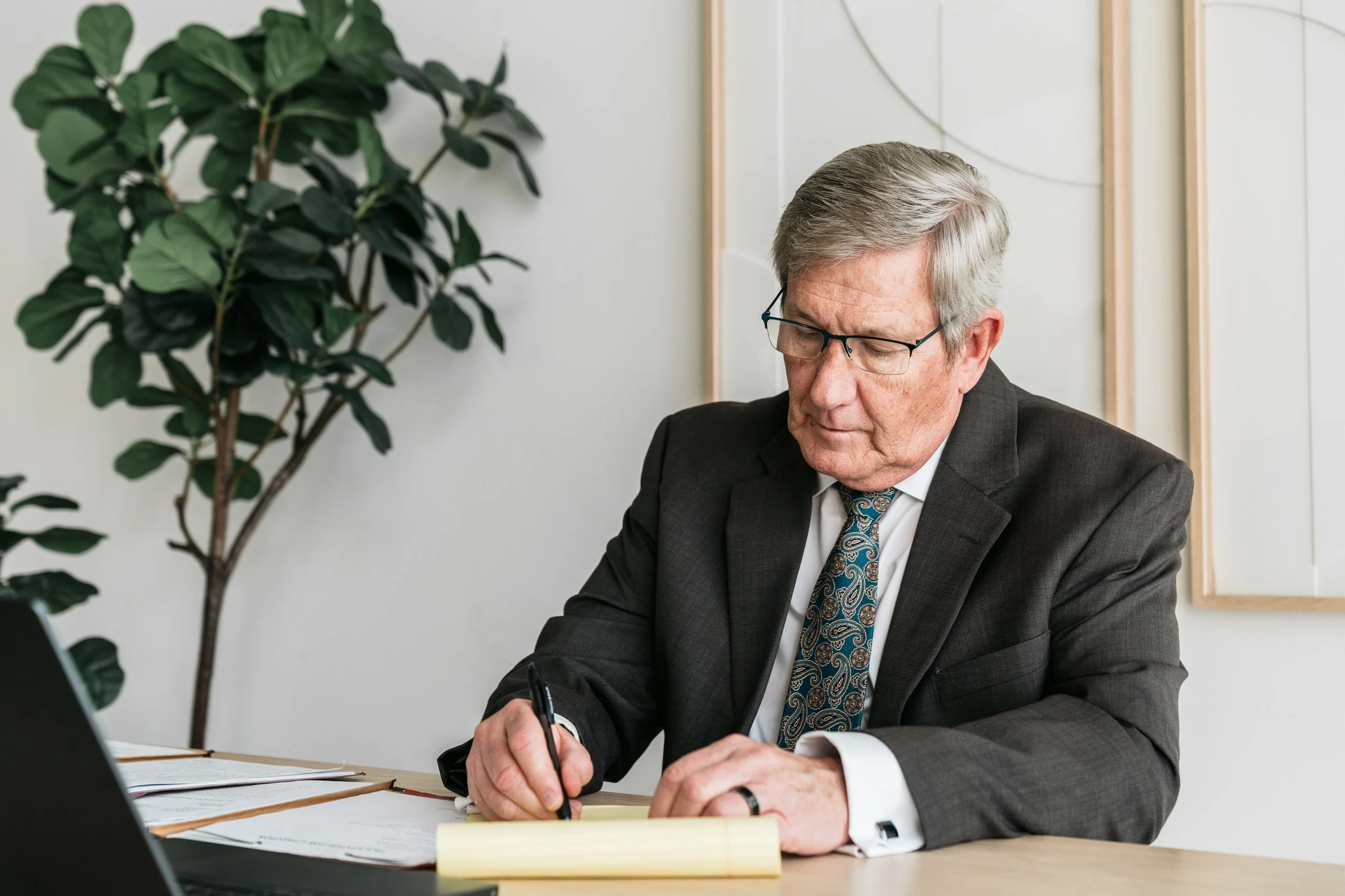 An older man wearing glasses and a suit, sitting at a desk, writing on a yellow notepad.