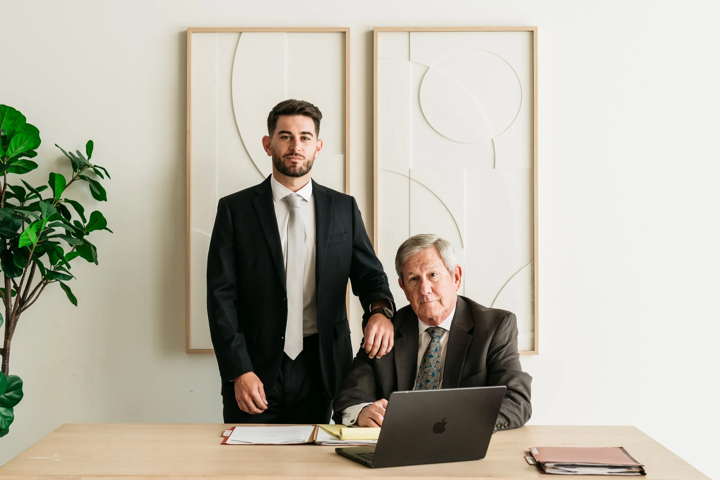 Two men in suits, one younger standing and one older sitting at a desk with a laptop, in a modern office with a minimalist wall art and green plant