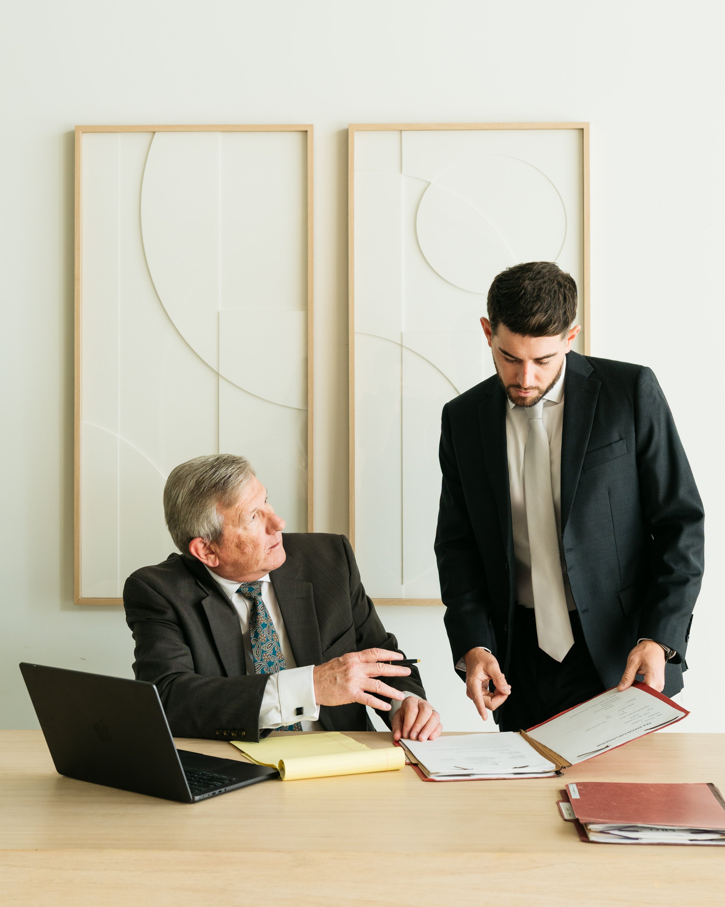 Two men in formal business attire having a discussion in an office. One is seated at a desk with a laptop and documents, the other is standing and reviewing papers. There are two decorative art pieces on the wall behind them.
