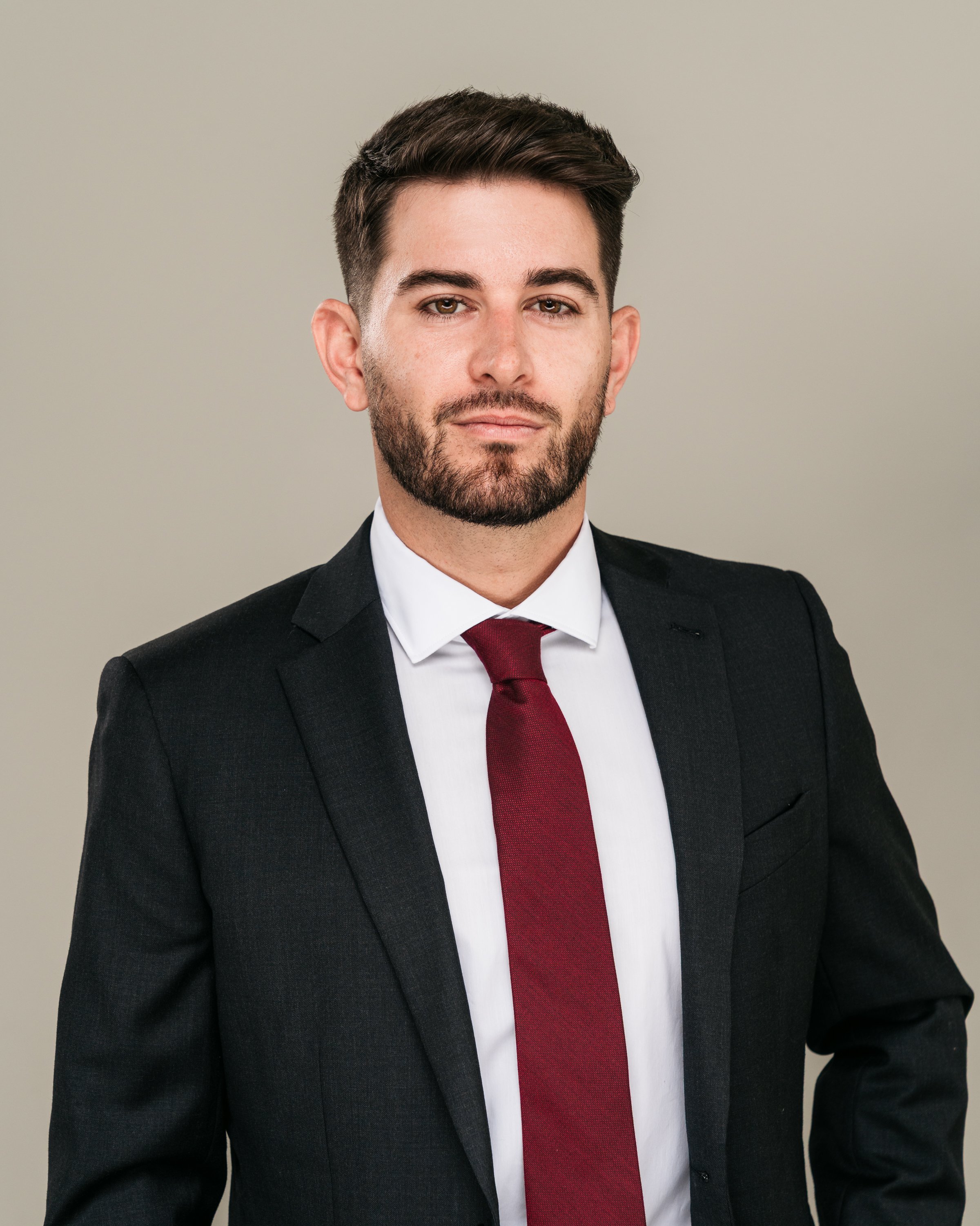 A man in a black suit, white shirt, and burgundy tie looking directly at the camera against a plain light gray background.
