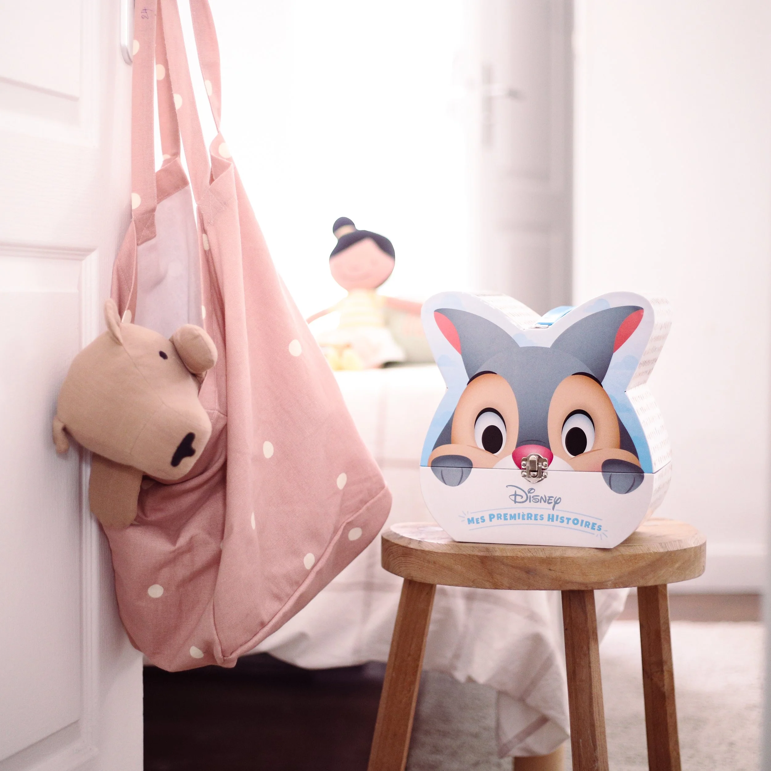 Une boîte à souvenir de Contes de Disney en forme de chat avec un visage de personnage Disney, placée sur un petit tabouret en bois. À gauche, un sac en tissu en forme d'ours accroché à une porte, et derrière, une poupée en arrière-plan dans une chambre lumineuse.