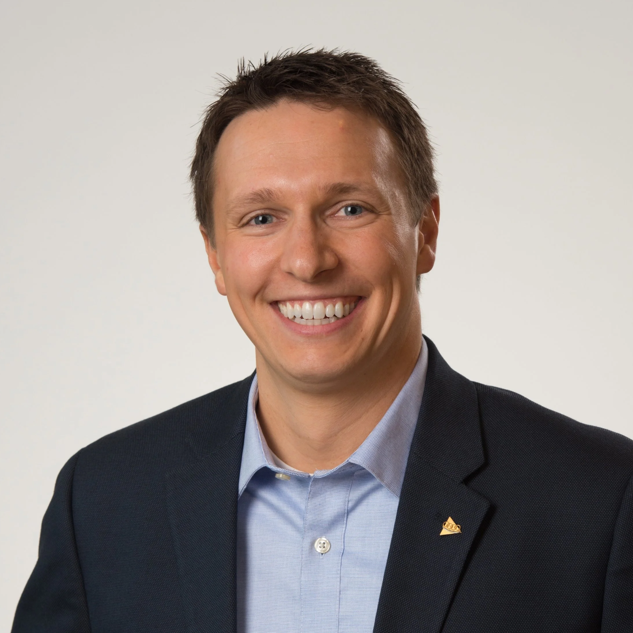 Close-up portrait of a man with light skin, short brown hair, smiling, dressed in a dark suit and light blue shirt, against a plain light background.
