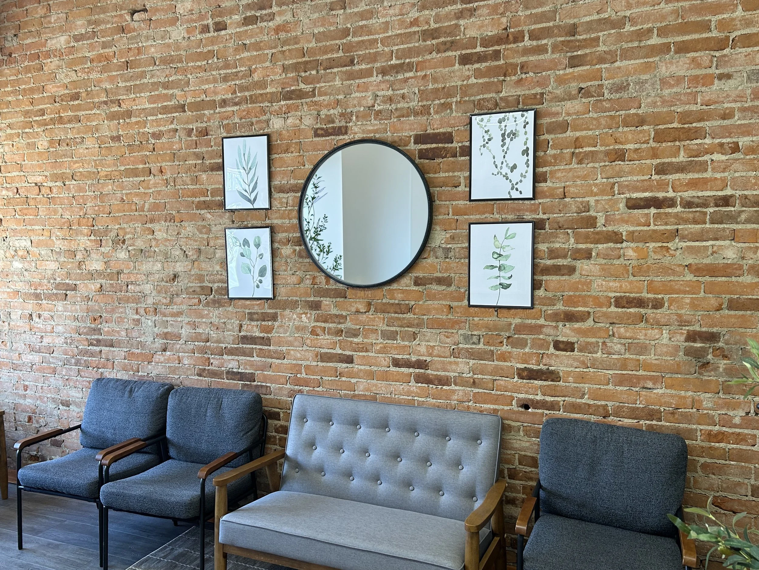 A seating area with four chairs in front of a brick wall decorated with four botanical prints and a round mirror, in a cozy indoor space.