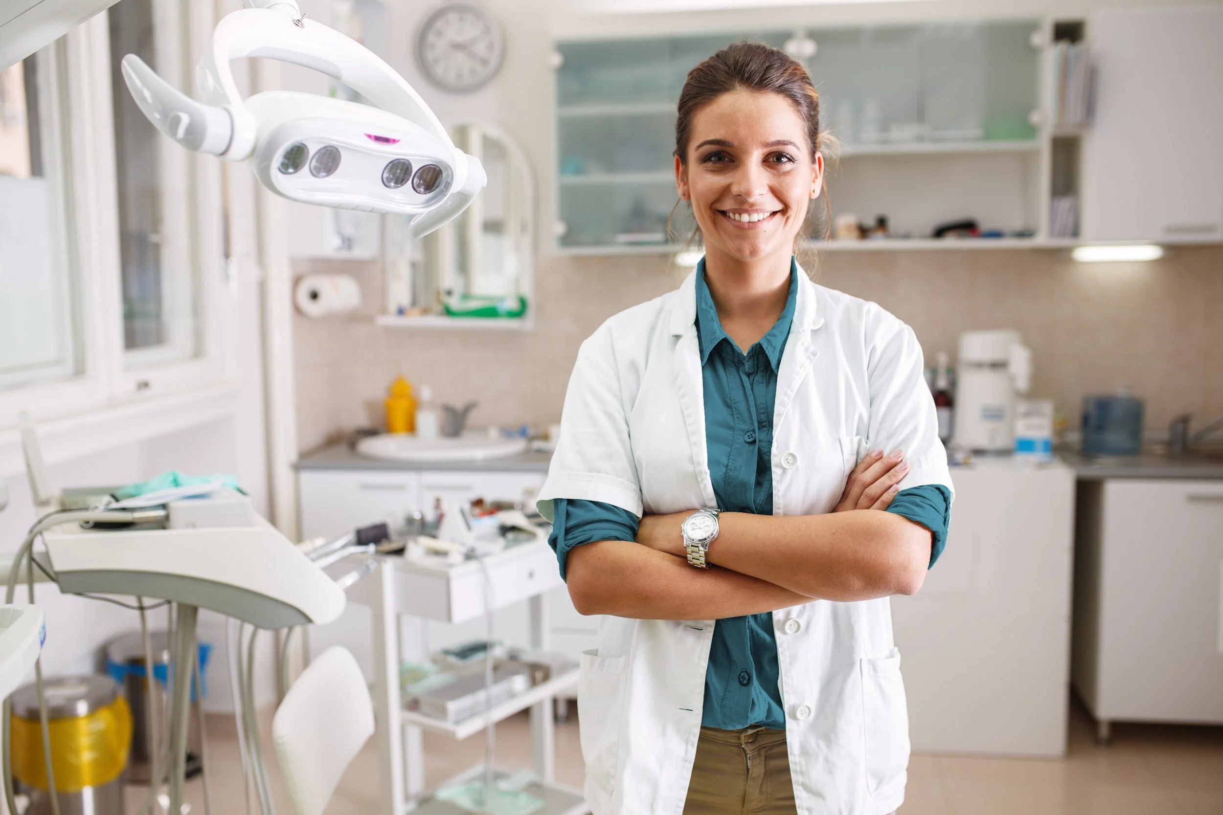 A smiling female healthcare professional standing with arms crossed in a medical or dental clinic. She is wearing a white coat, a teal shirt, and a watch. Medical equipment and furniture are visible in the background.