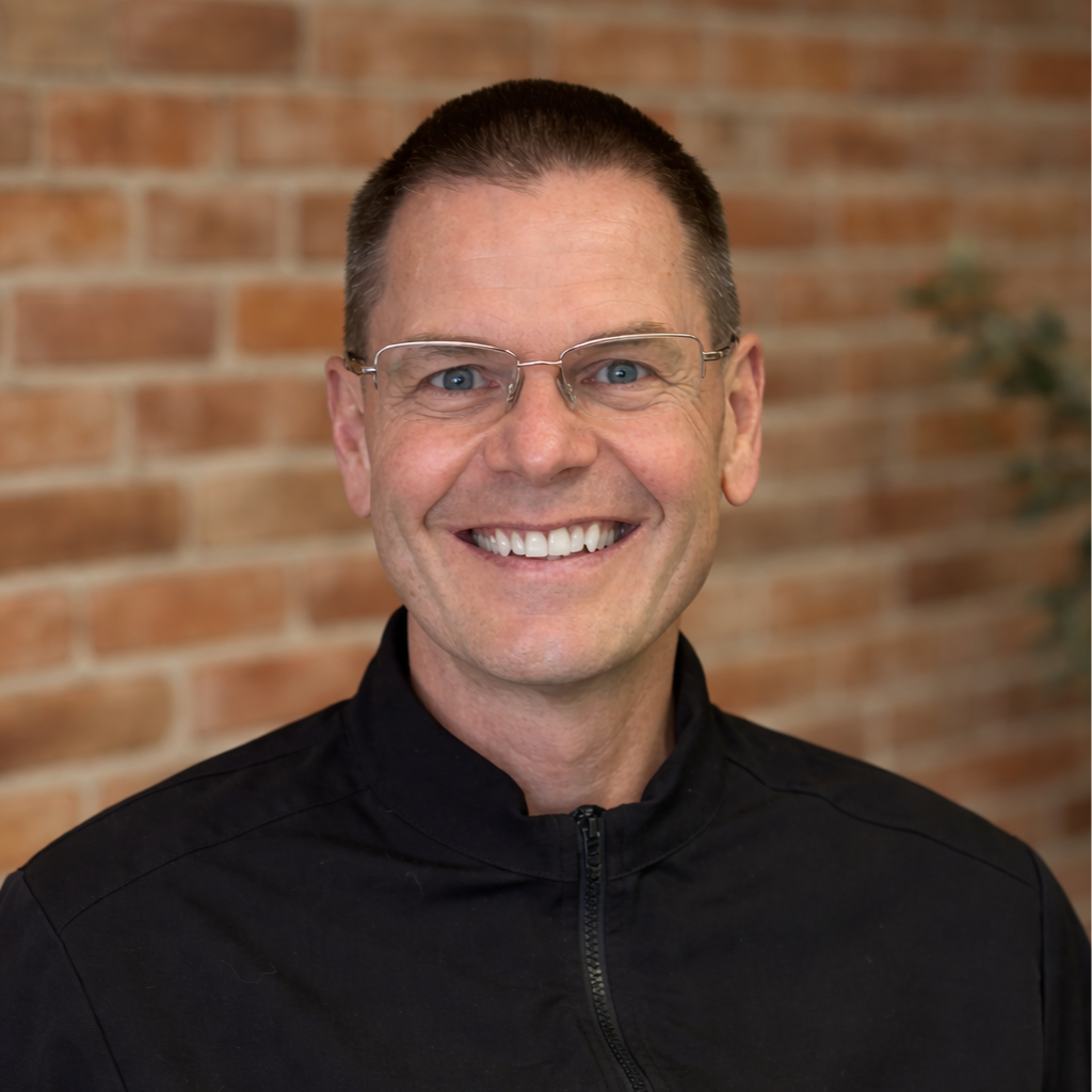 A smiling man with short brown hair, glasses, and a black zip-up jacket stands in front of a brick wall.