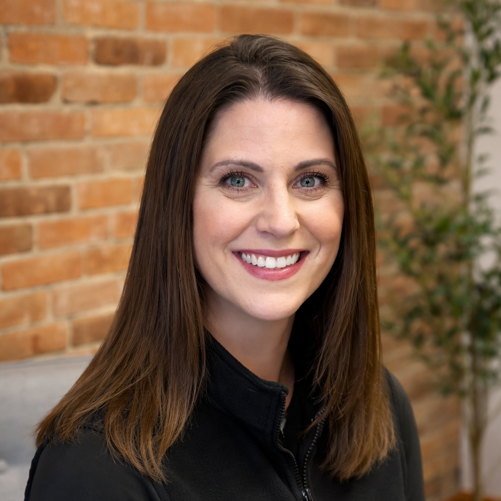 A woman with straight brown hair, blue eyes, and a bright smile, wearing a black zip-up top, in front of a brick wall with a potted plant in the background.