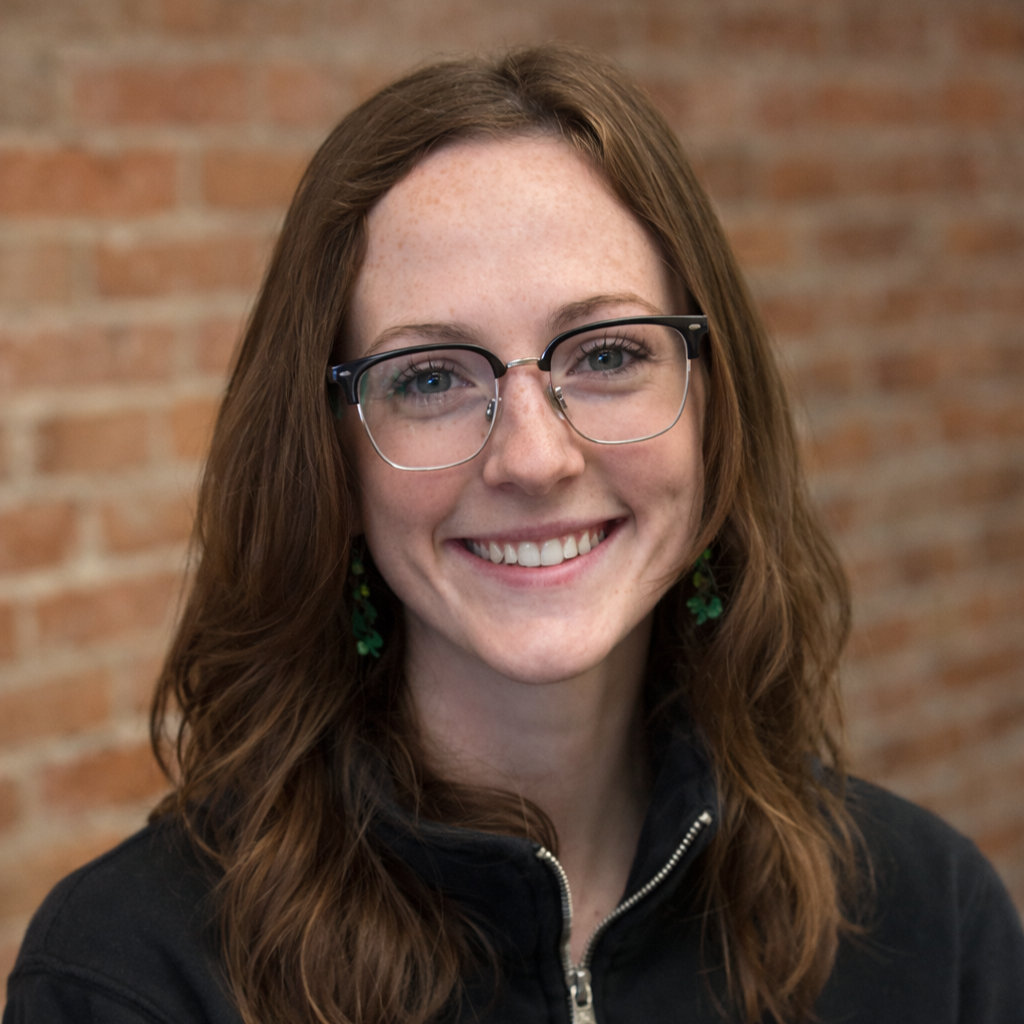 A young woman with long, wavy red hair and glasses, smiling, in front of a brick wall.