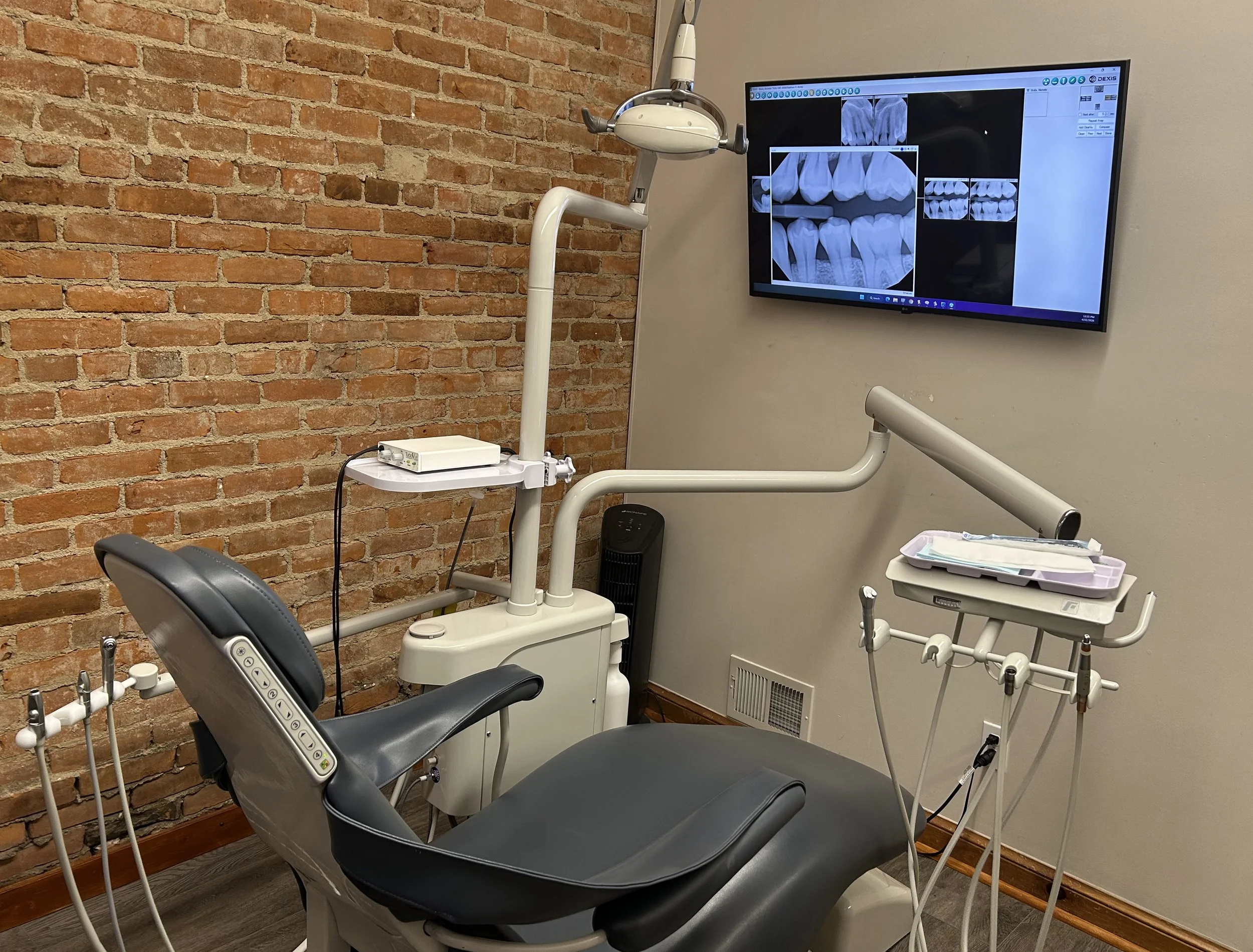 A dental examination room with a brick wall, dental chair, dental light, and monitor displaying dental X-ray images of teeth.