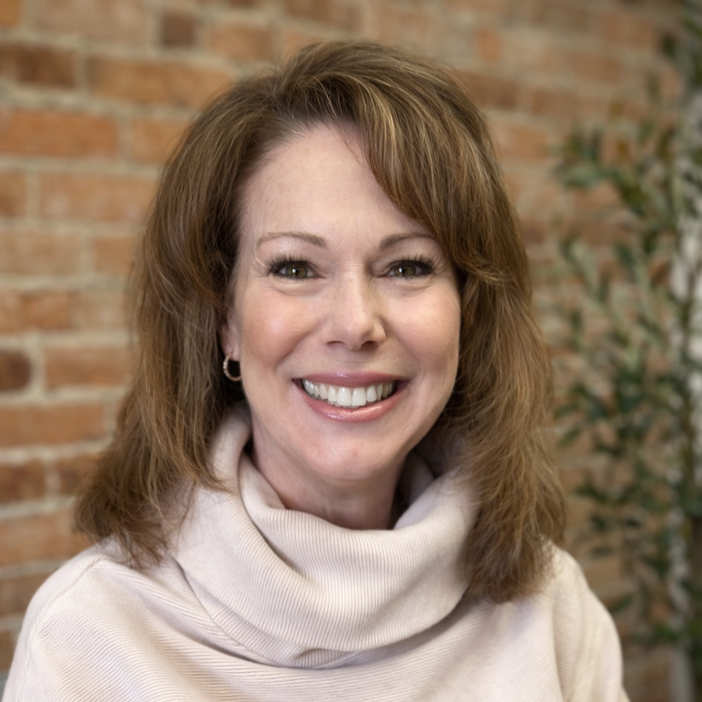 A woman with shoulder-length brown hair wearing a beige turtleneck sweater, smiling in front of a brick wall with some greenery in the background.