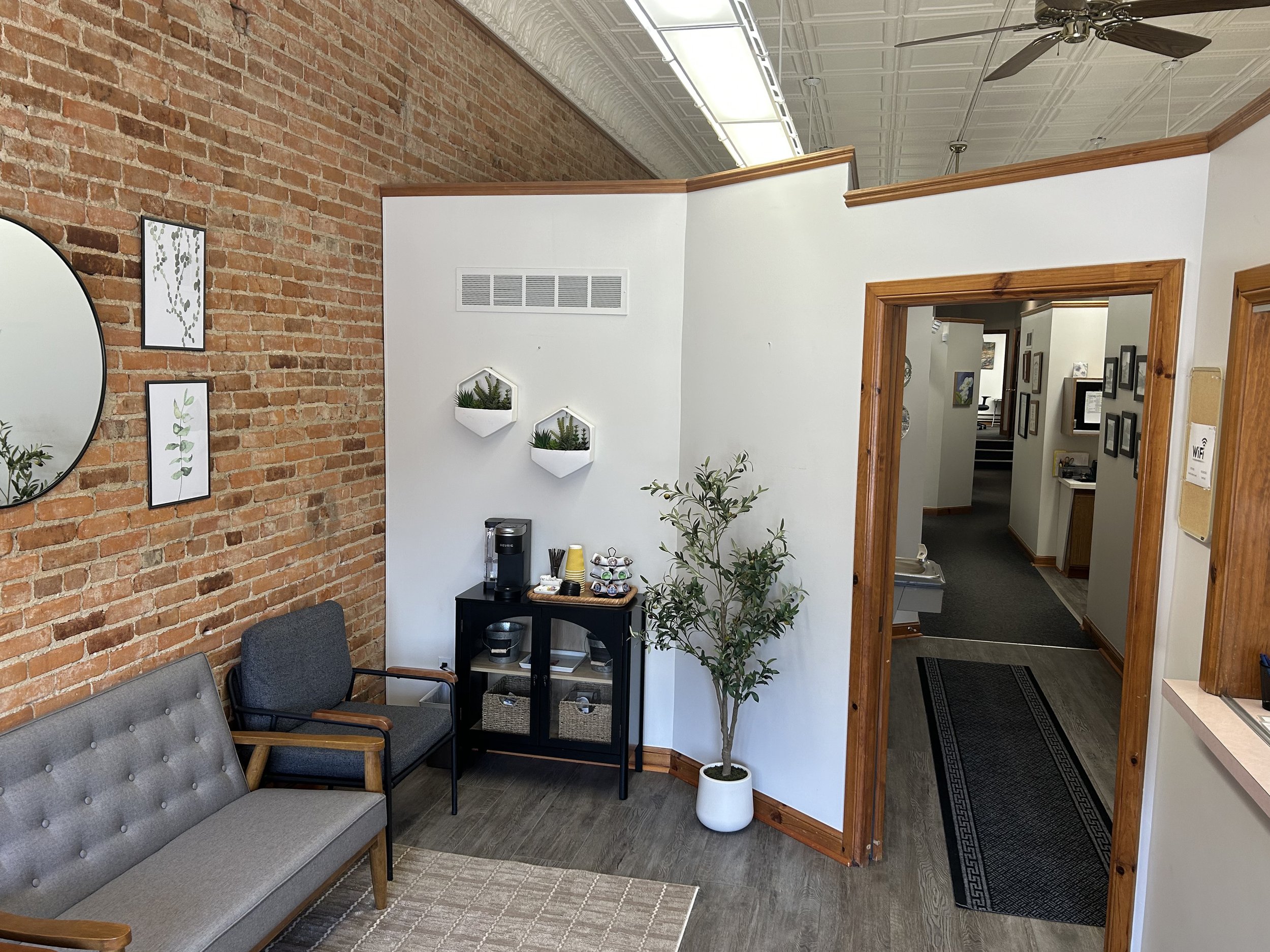 A waiting area with a gray mid-century modern style sofa, two gray chairs with wooden arms, a small black table with jute storage baskets and a coffee station, a tall potted plant, and a white wall with three wall-mounted planters containing succulents. The brick wall and an open doorway to a hallway are visible.
