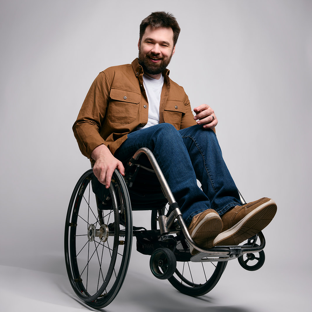 John Loeppky, host of History in 60, seated in his wheelchair and smiling, wearing a brown shirt over a white tshirt, jeans & brown shoes