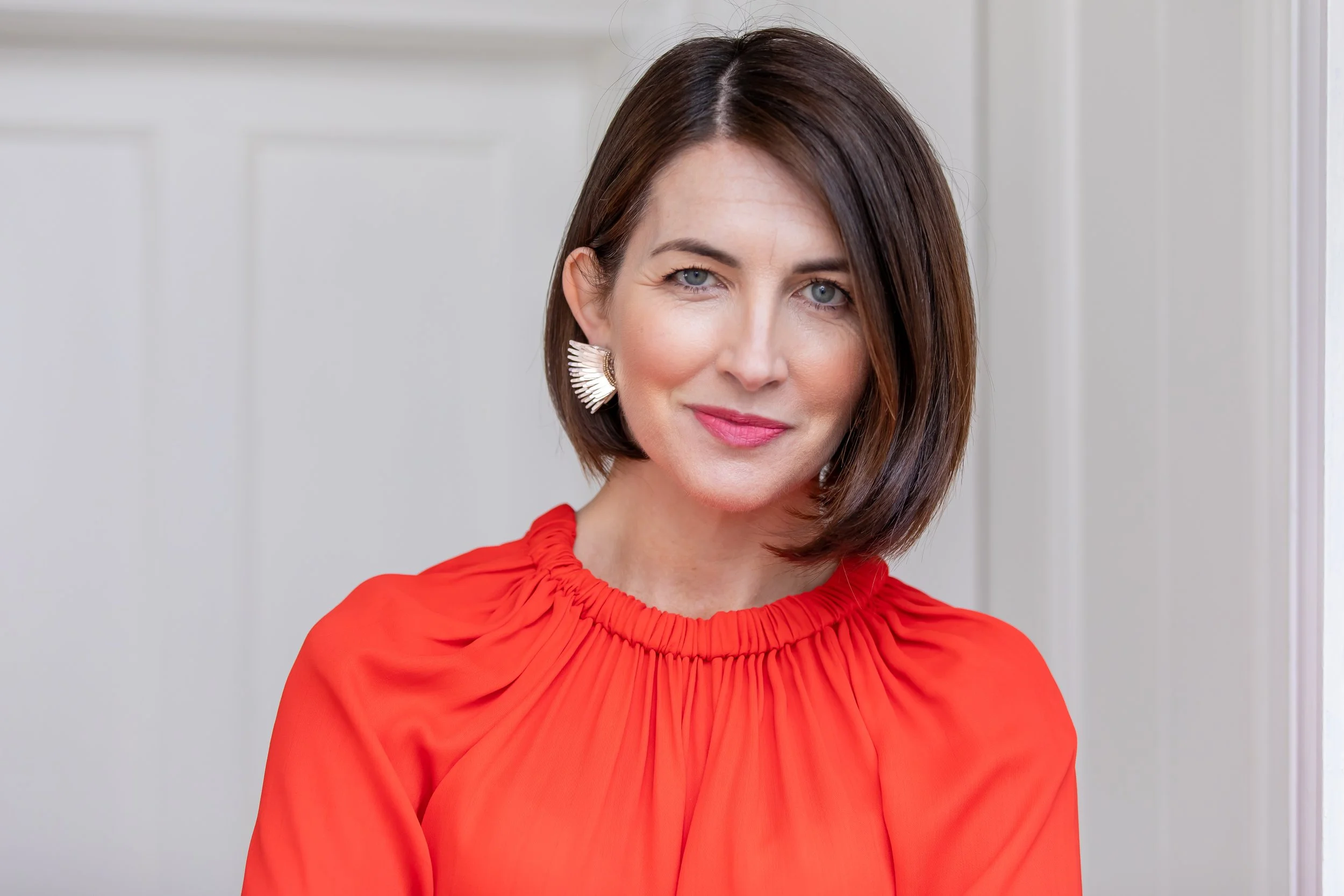 A woman with short brown hair wearing a red blouse with pleated details at the neckline and large dangling earrings, standing indoors with a white wall background.