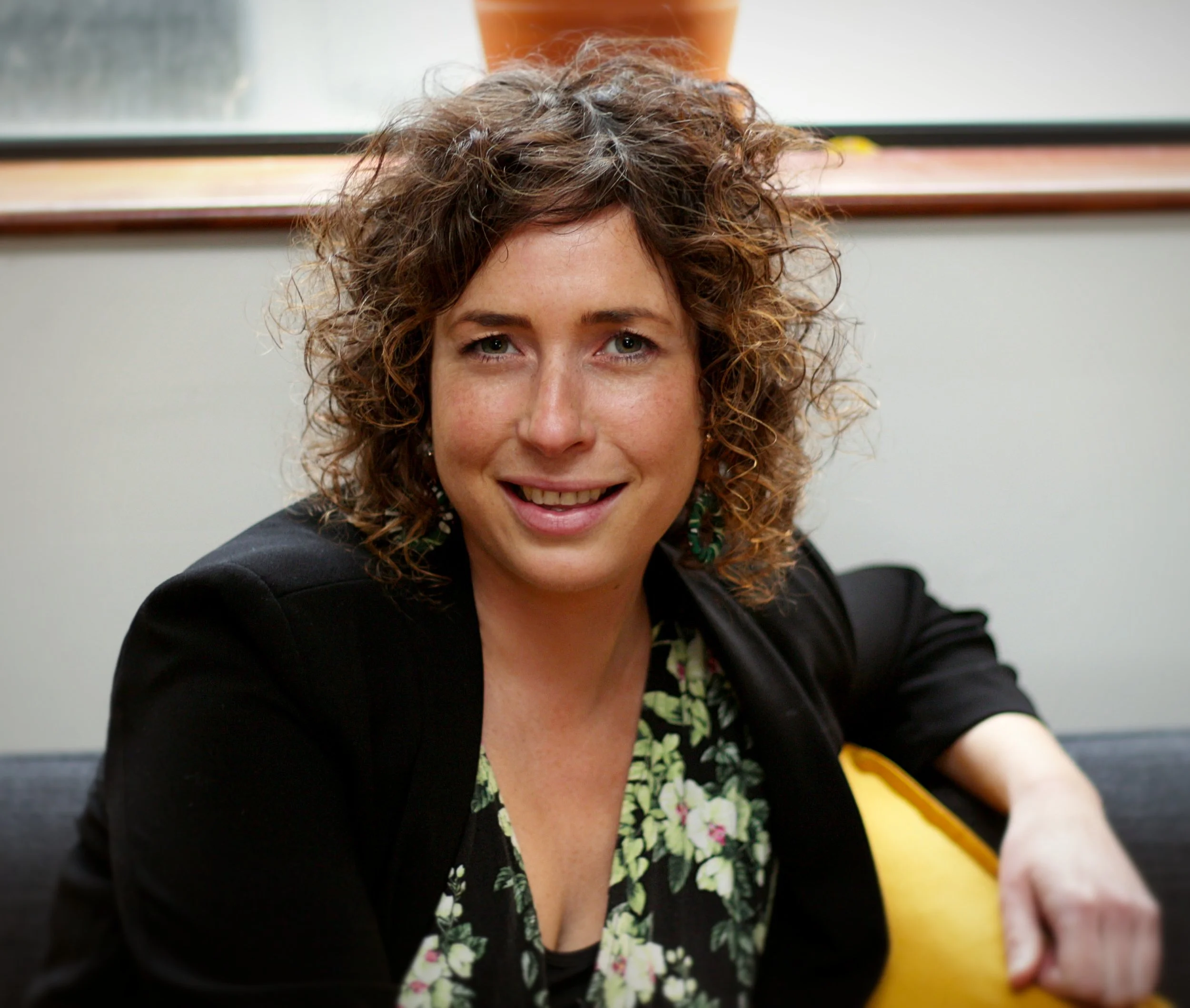 A woman with curly brown hair wearing a black blazer and floral top sitting near a window with a potted plant and a yellow object behind her.