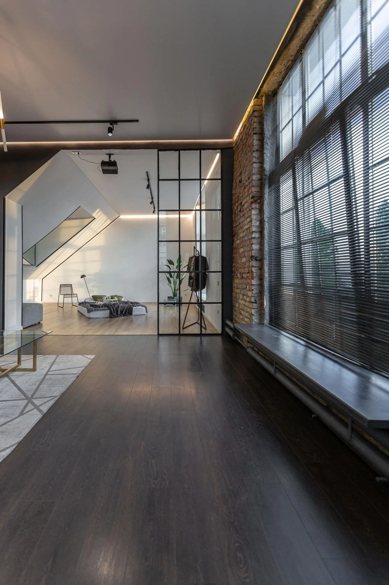 Modern interior space with dark hardwood floors, large windows with blinds, exposed brick wall, glass partition, minimalist furniture, and decorative lighting elements.