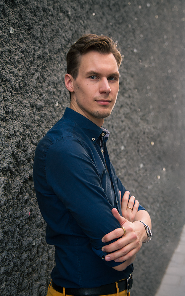 A young man with short, light brown hair and blue eyes standing against a dark, textured wall. He's wearing a dark blue button-up shirt with the sleeves rolled up, yellow pants, a wristwatch, and a ring on his finger. He has a confident expression with arms crossed.