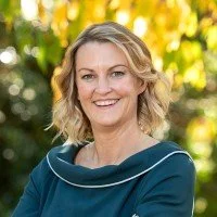 A smiling woman with blonde wavy hair standing outdoors in front of greenery and yellow flowers.