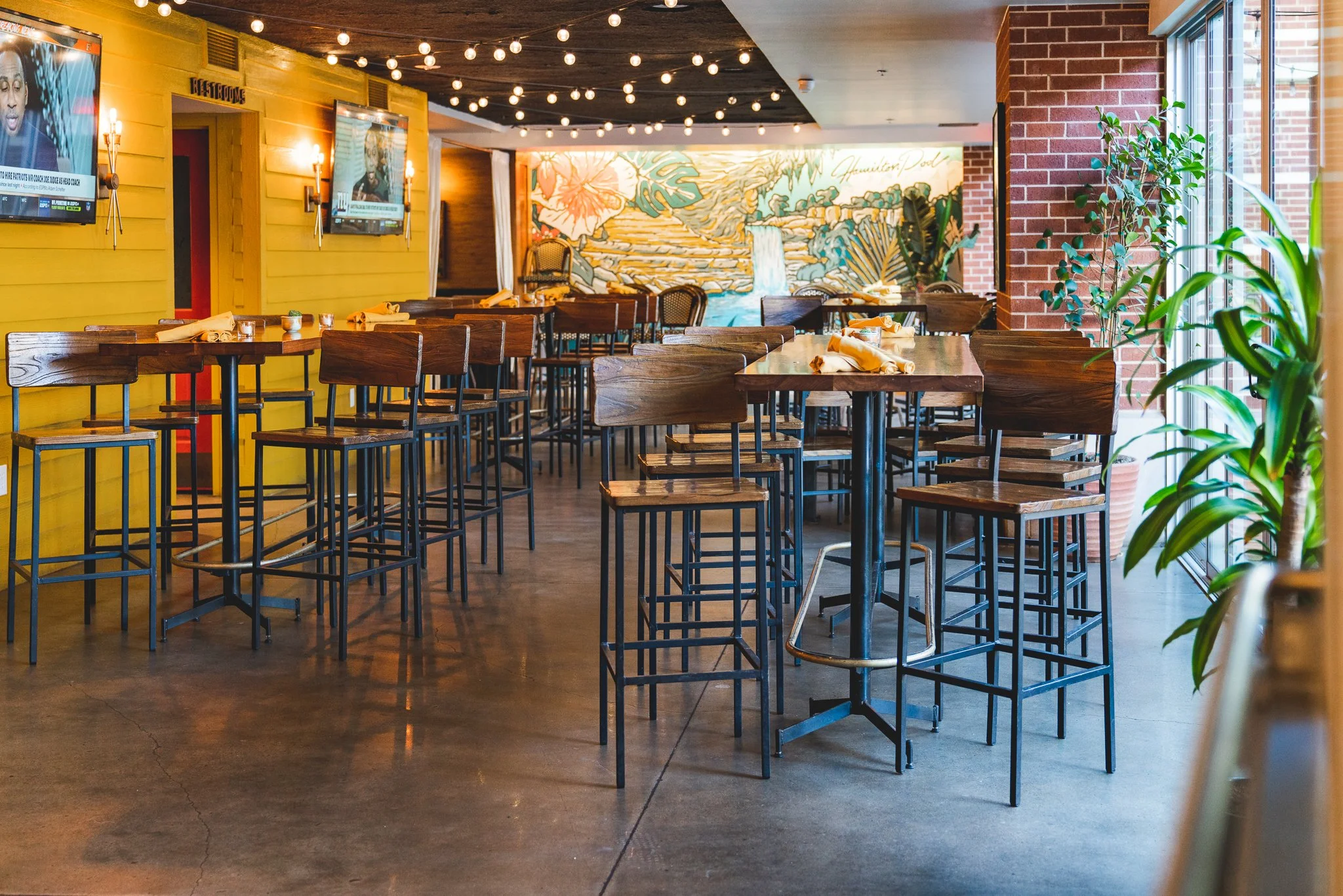 Empty restaurant with high wooden tables and chairs, decorated with potted plants, yellow and brick walls, large windows, and a mural of a waterfall scene.