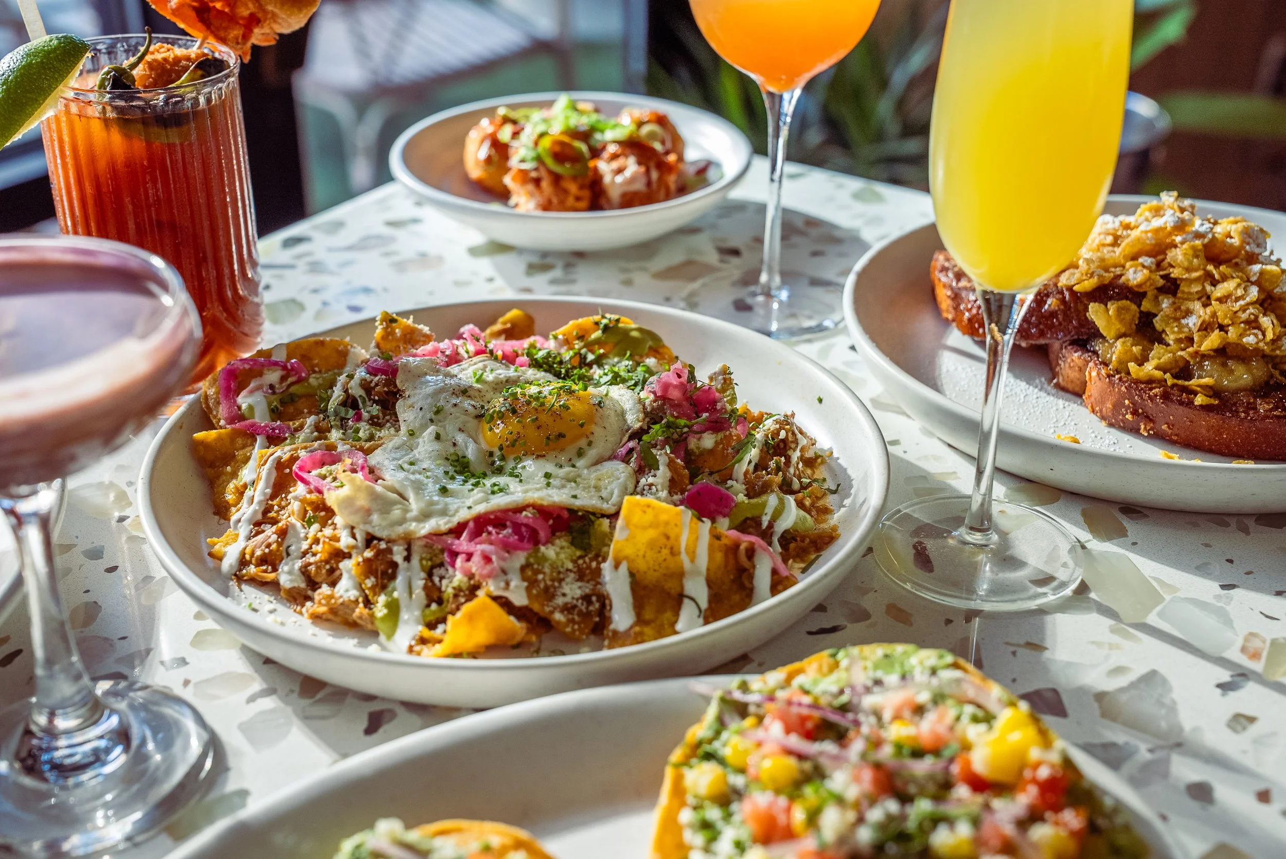 A table filled with several dishes and drinks, including a plate of nachos topped with a fried egg, pink onions, and sauces, various margaritas and cocktails, and slices of toast with toppings in a bright, casual setting.