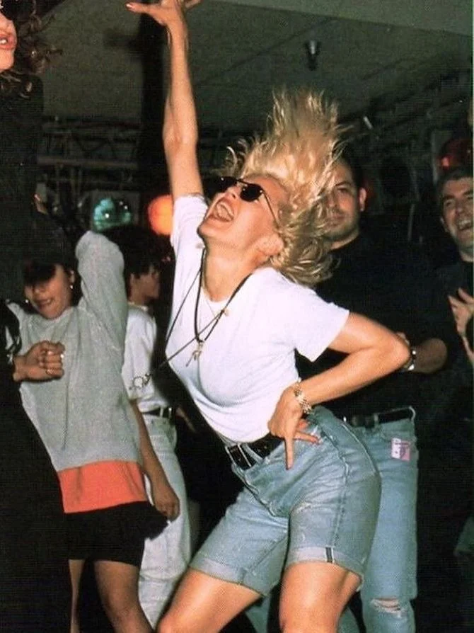 A woman dancing exuberantly at a party or club, with her hair flying and wearing sunglasses, a white t-shirt, and denim shorts. She looks joyful and energetic among other smiling people.