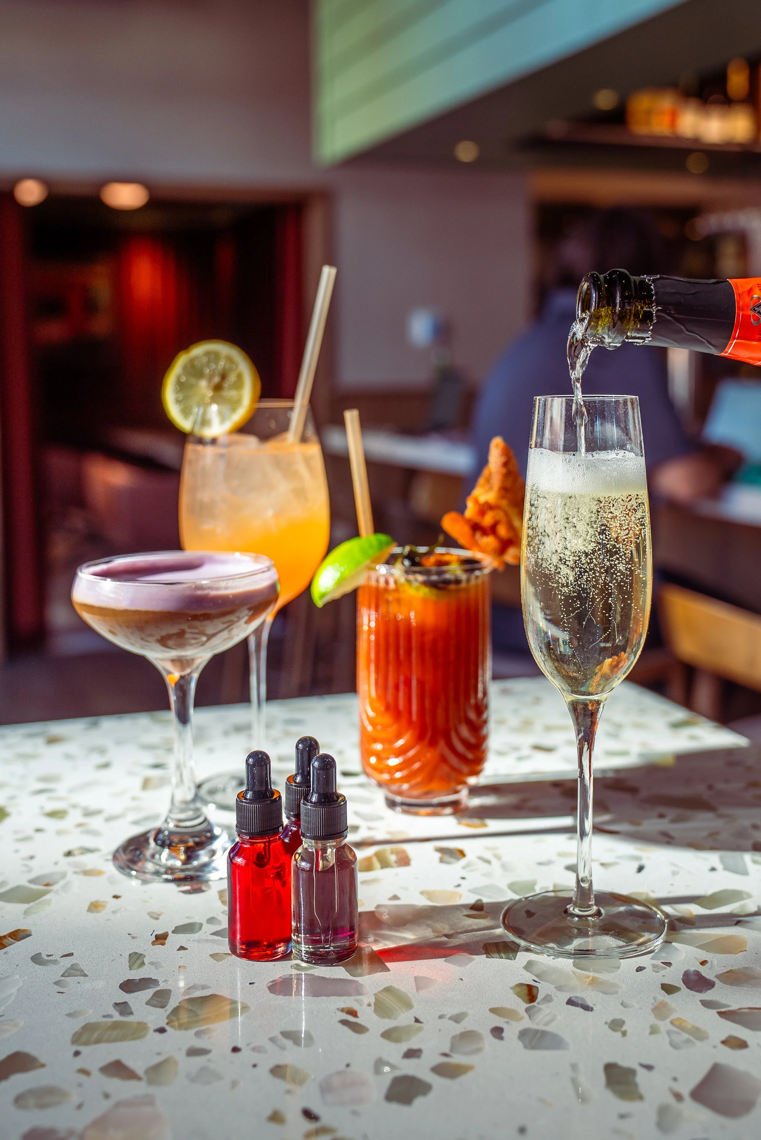 A table with four cocktails and three small bottles of flavoring in a restaurant bar setting.