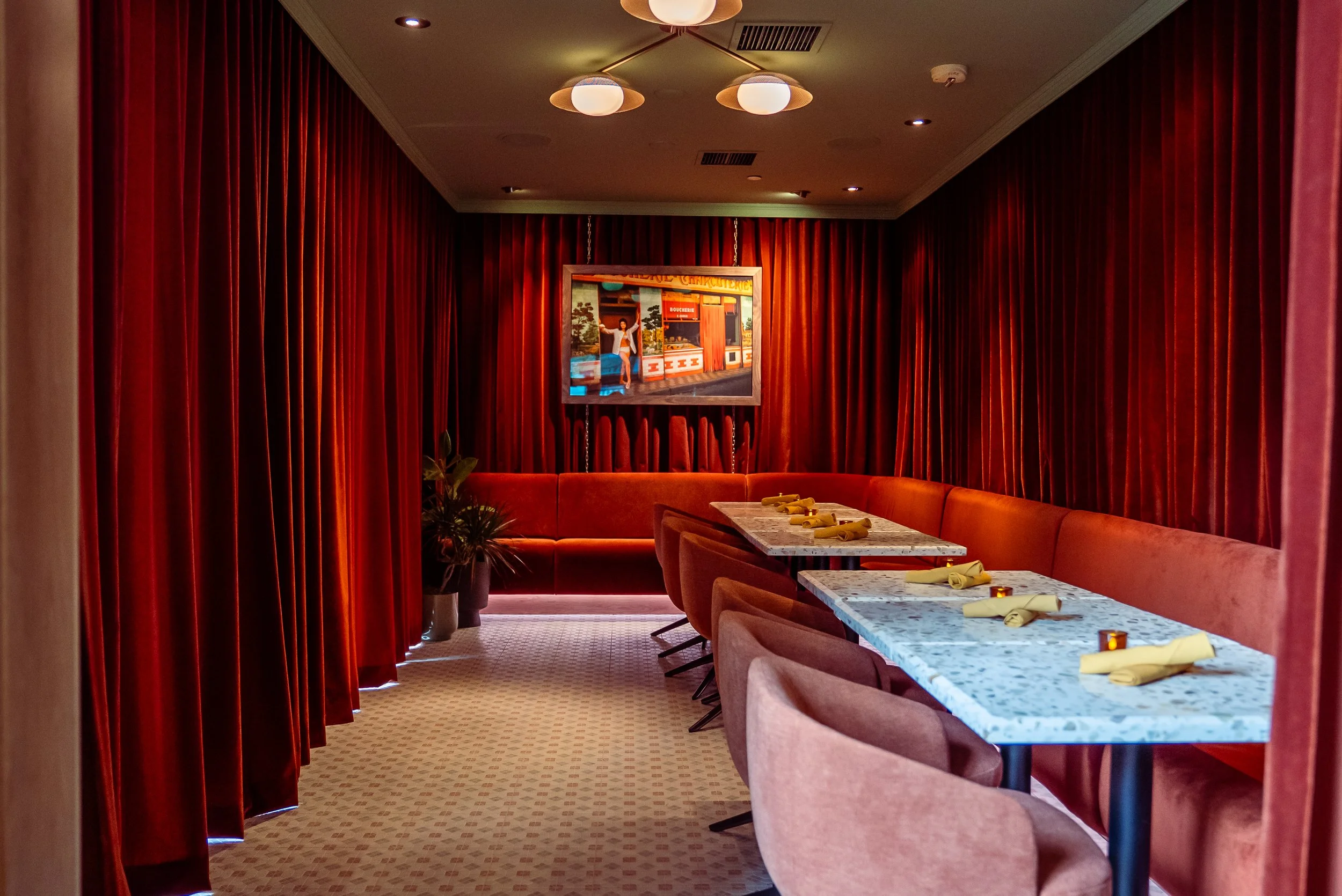 A private dining room with red velvet curtains, a long marble table with napkins and candles, pink upholstered chairs, a red velvet bench, and a framed picture hanging on the back wall.