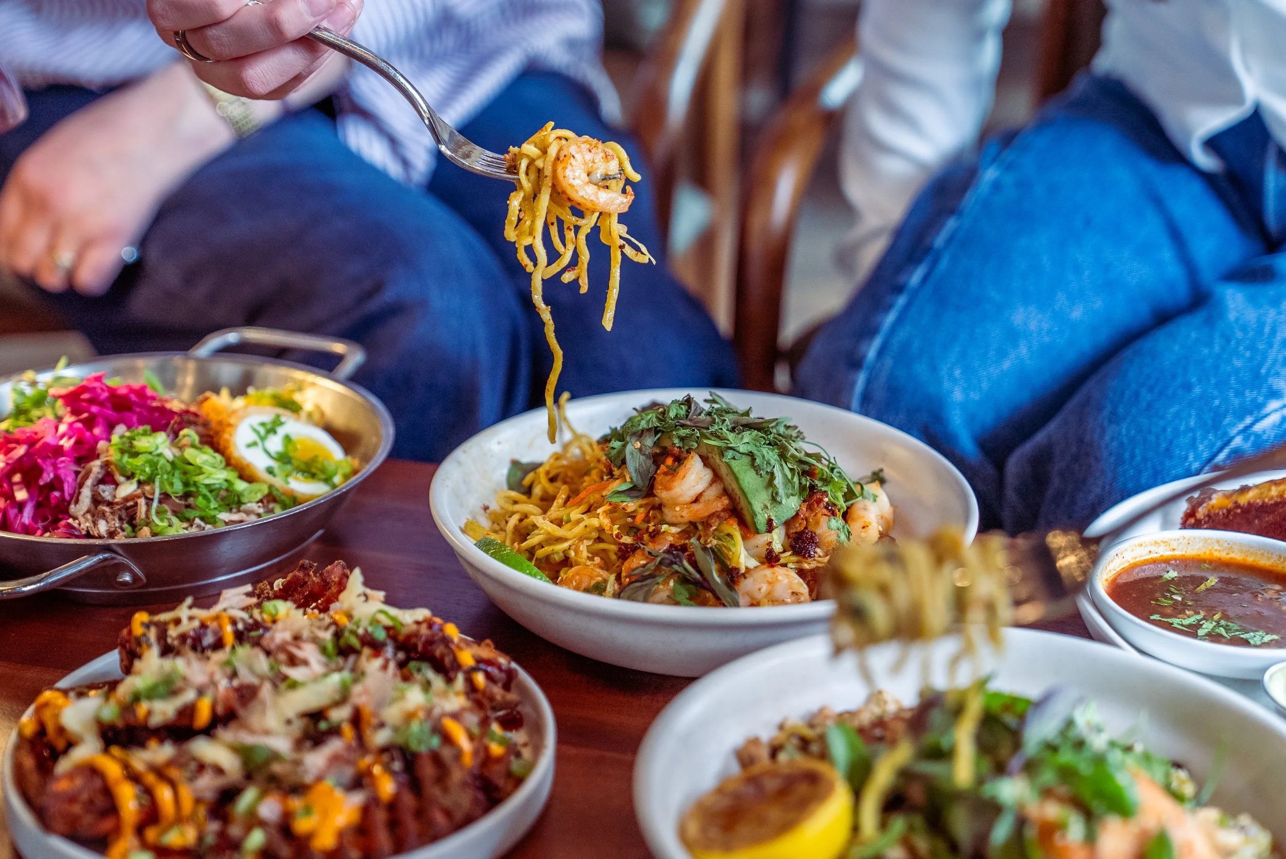 A person using a fork to pick up noodles with shrimp from a bowl of Asian cuisine, surrounded by various dishes including salads, bowls of soup, and other Asian dishes on a wooden table.