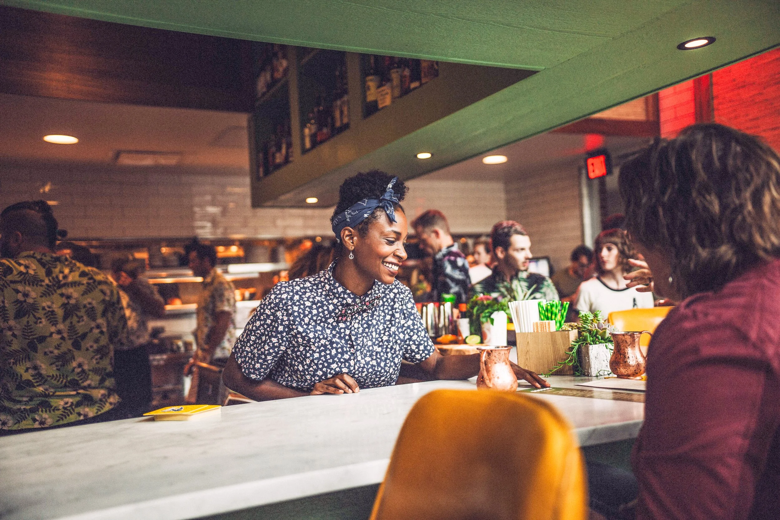 A woman working behind the bar counter smiling at a customer in a lively bar or restaurant with other patrons in the background.