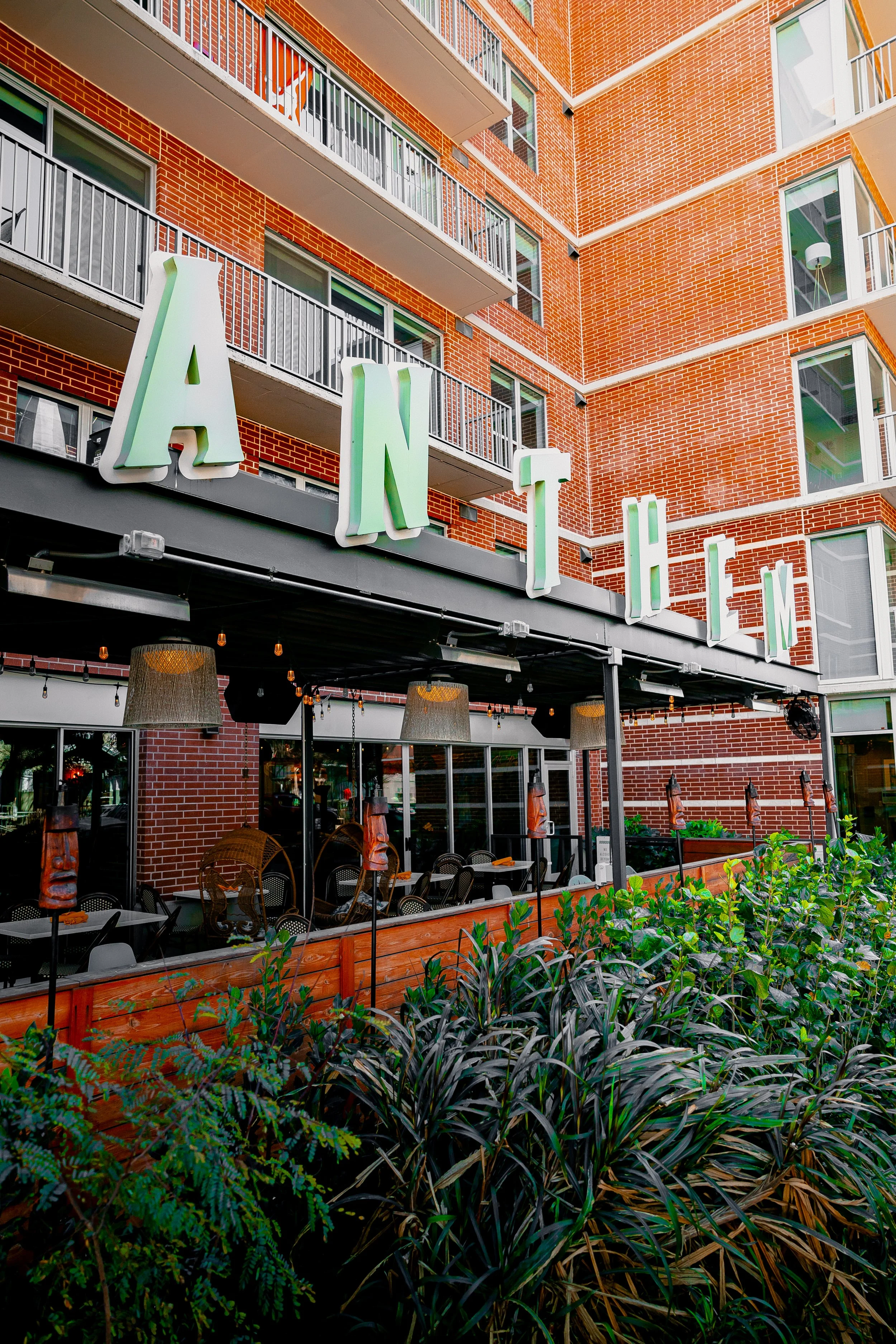 Exterior view of a restaurant named 'ANTHEM' with a brick building and outdoor seating area, plants in the foreground, and large illuminated letters spelling 'ANTHEM' on the building.