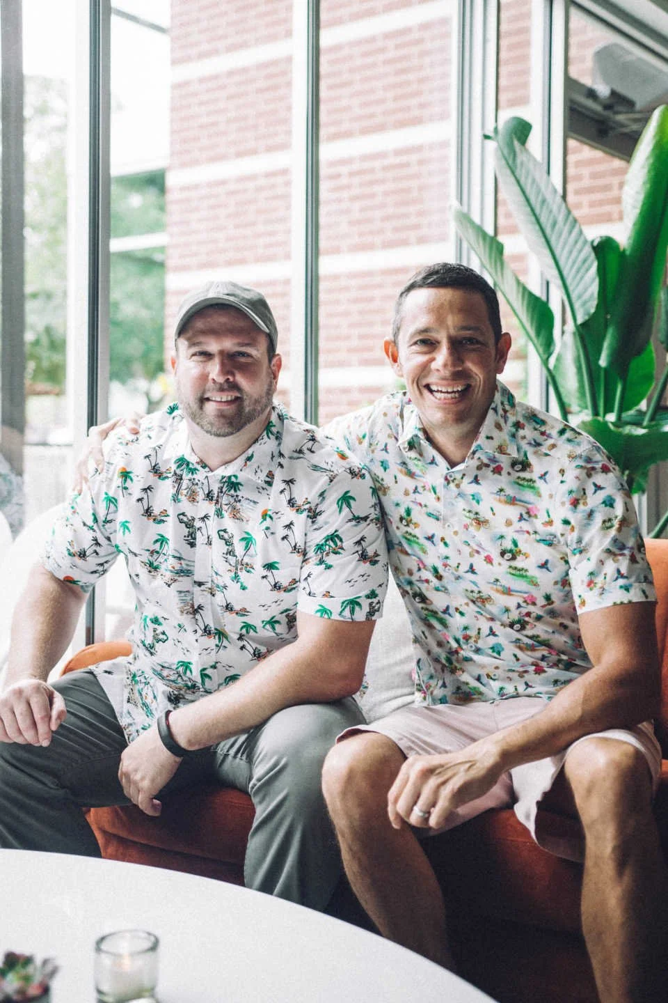 Two men sitting close together, smiling, wearing casual Hawaiian shirts, indoors with large plants and a large window behind them.
